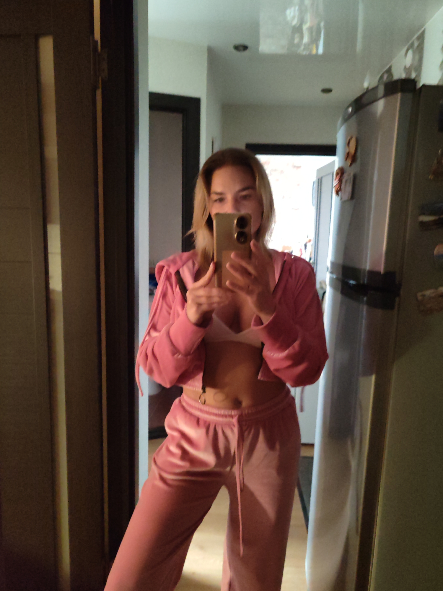 A fair-skinned woman is taking a selfie in front of a mirror. She is wearing a pink tracksuit and a white bra. The background shows a doorway, a refrigerator, and a glimpse of a room through the doorway. The location appears to be indoors. There's no information in the image suggesting the city of Petropavlovsk-Kamchatsky, Russia.