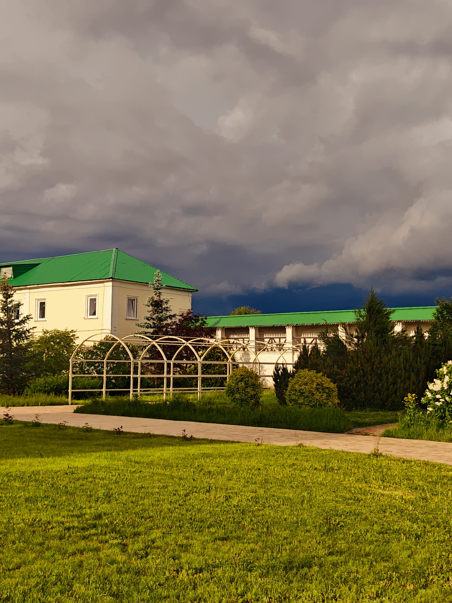 The image depicts a building with a green roof in Луговой, Russia. The building is surrounded by green grass and trees, and a white metal structure resembling an archway is in the foreground. The sky is filled with dark, ominous clouds, suggesting an approaching storm. No people are visible in the scene.
