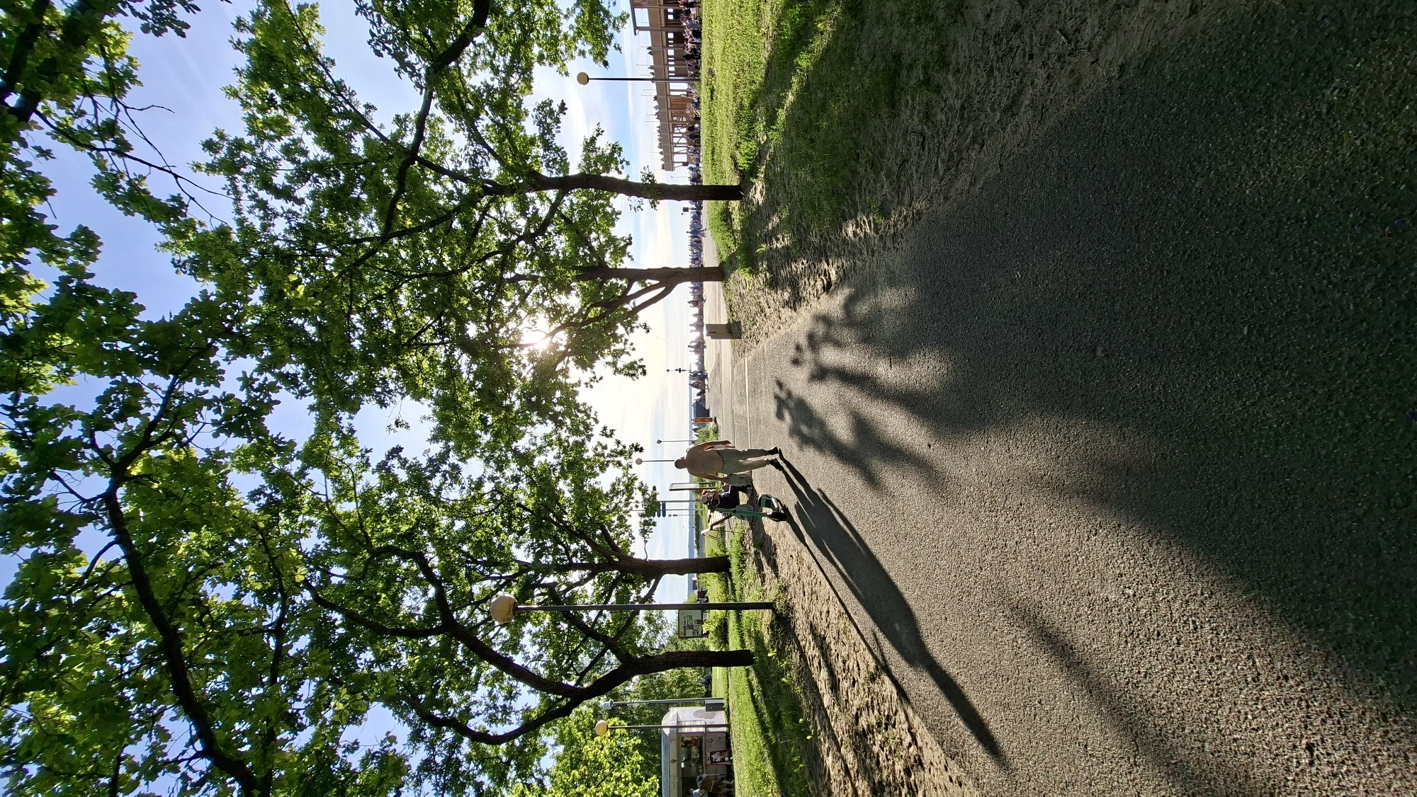 A man walks along a paved path in Tallinn, Estonia, with a scooter beside him. The path is lined with trees casting long shadows. In the background, a body of water is visible, with buildings and people along the shore. The sun shines through the leaves of the trees, creating a dappled light effect.