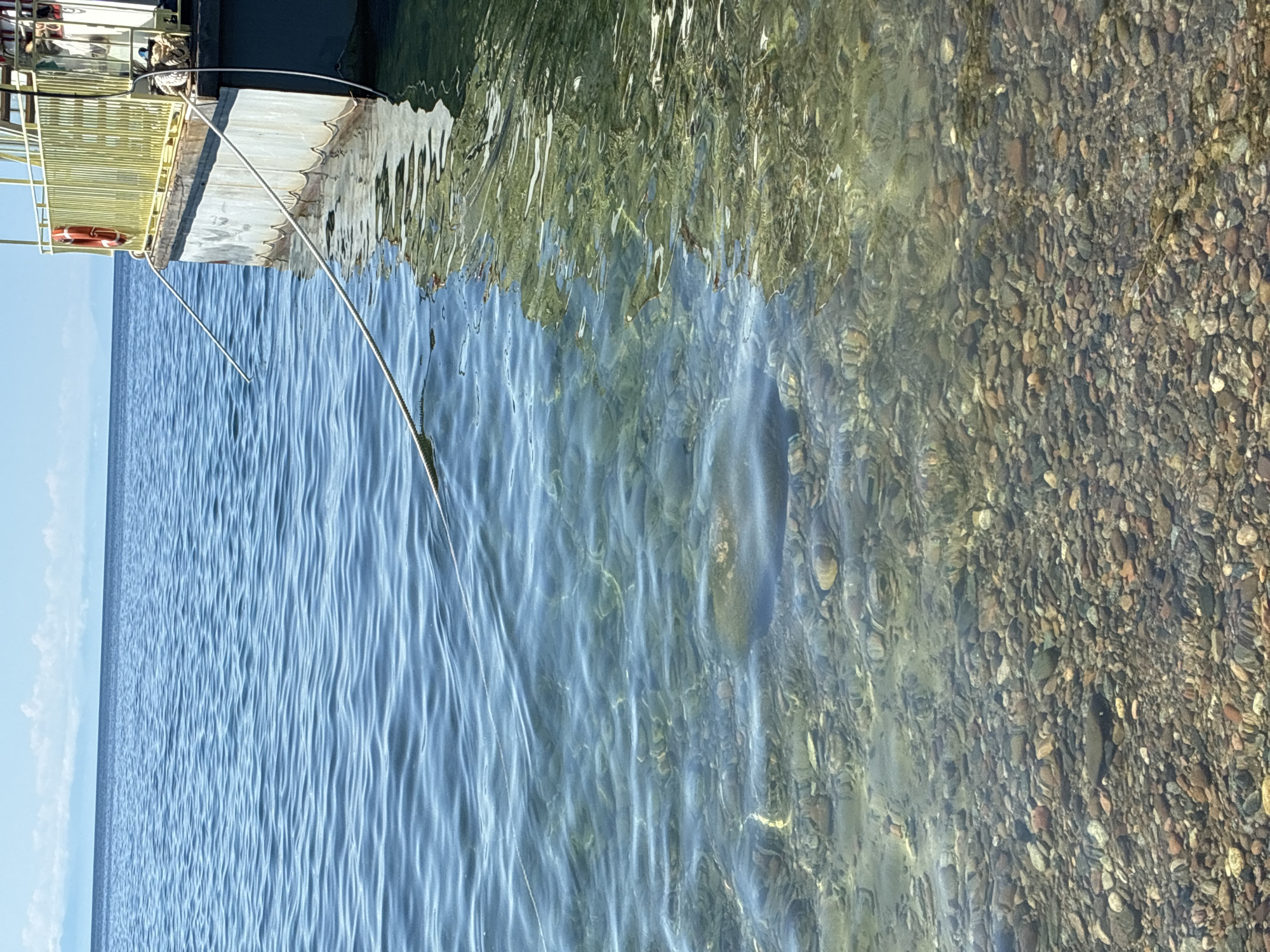 The image depicts a clear, shallow body of water with a rocky bottom, likely the shore of Lake Baikal in Listvyanka, Russia. A boat, possibly a ferry, is docked on the right side of the frame. The boat has a yellow railing and a life preserver. Ropes connect the boat to the shore. The water is calm with gentle ripples, and sunlight creates reflections on the surface. The rocky seabed is visible through the transparent water. In the distance, a hazy shoreline or landmass can be seen under a bright, partly cloudy sky. No people are visible in the frame.