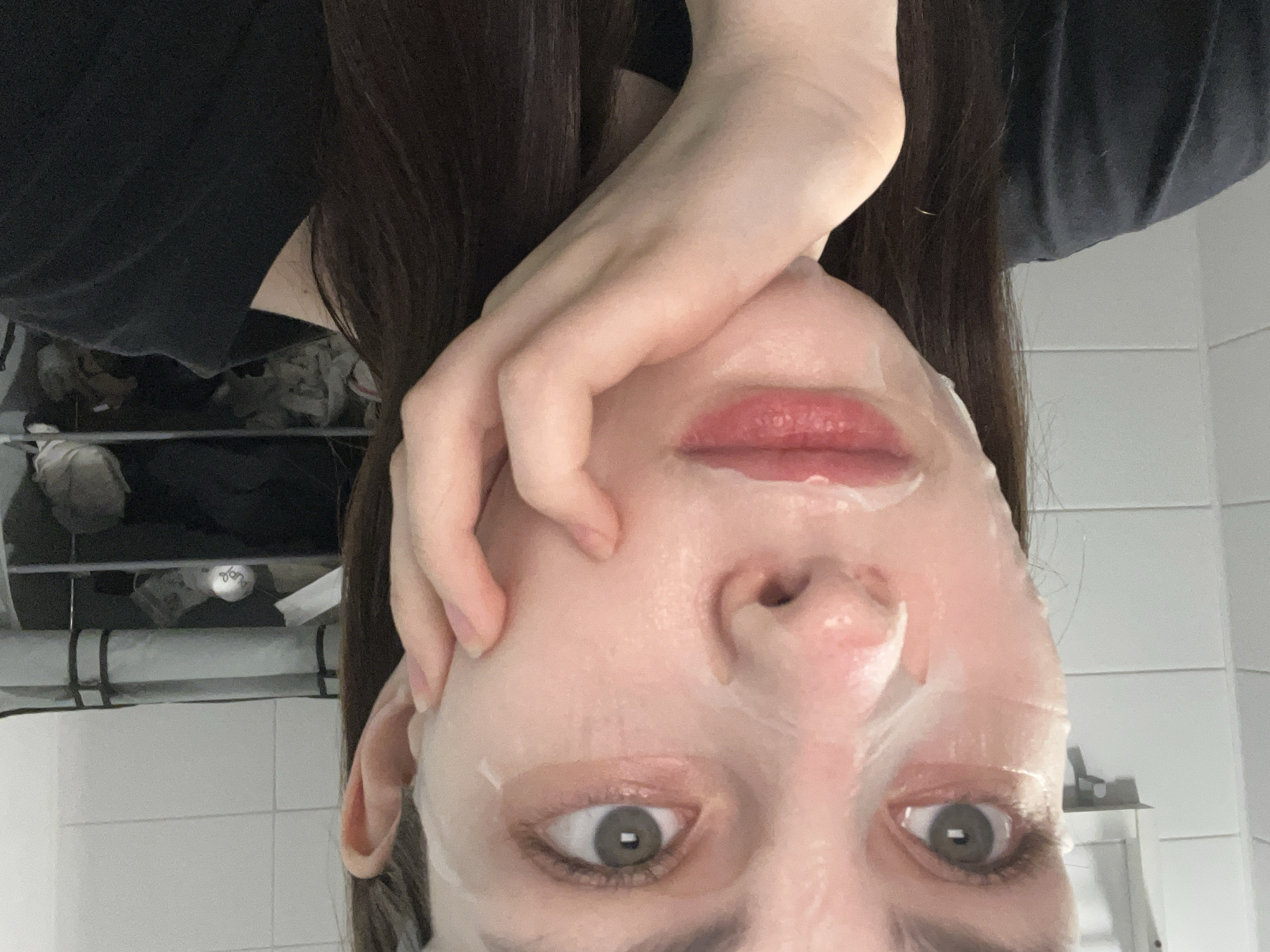The image presents a close-up, chest-up view of a female individual in an indoor residential setting in Saint Petersburg, Russia. The subject has fair skin, light-colored eyes, and dark brown hair, and is looking directly at the camera. A translucent sheet mask covers her face, leaving her eyes and mouth exposed. Her lips are a natural reddish-pink. Her right hand is positioned with fingers curled, resting against her jawline. She is wearing a dark-colored, possibly black, t-shirt. The background features light-colored tiled walls, indicative of a bathroom or similar utility area. To the right, a dark multi-shelf unit containing various indistinct objects is visible. A white wall-mounted fixture is partially visible on the left.