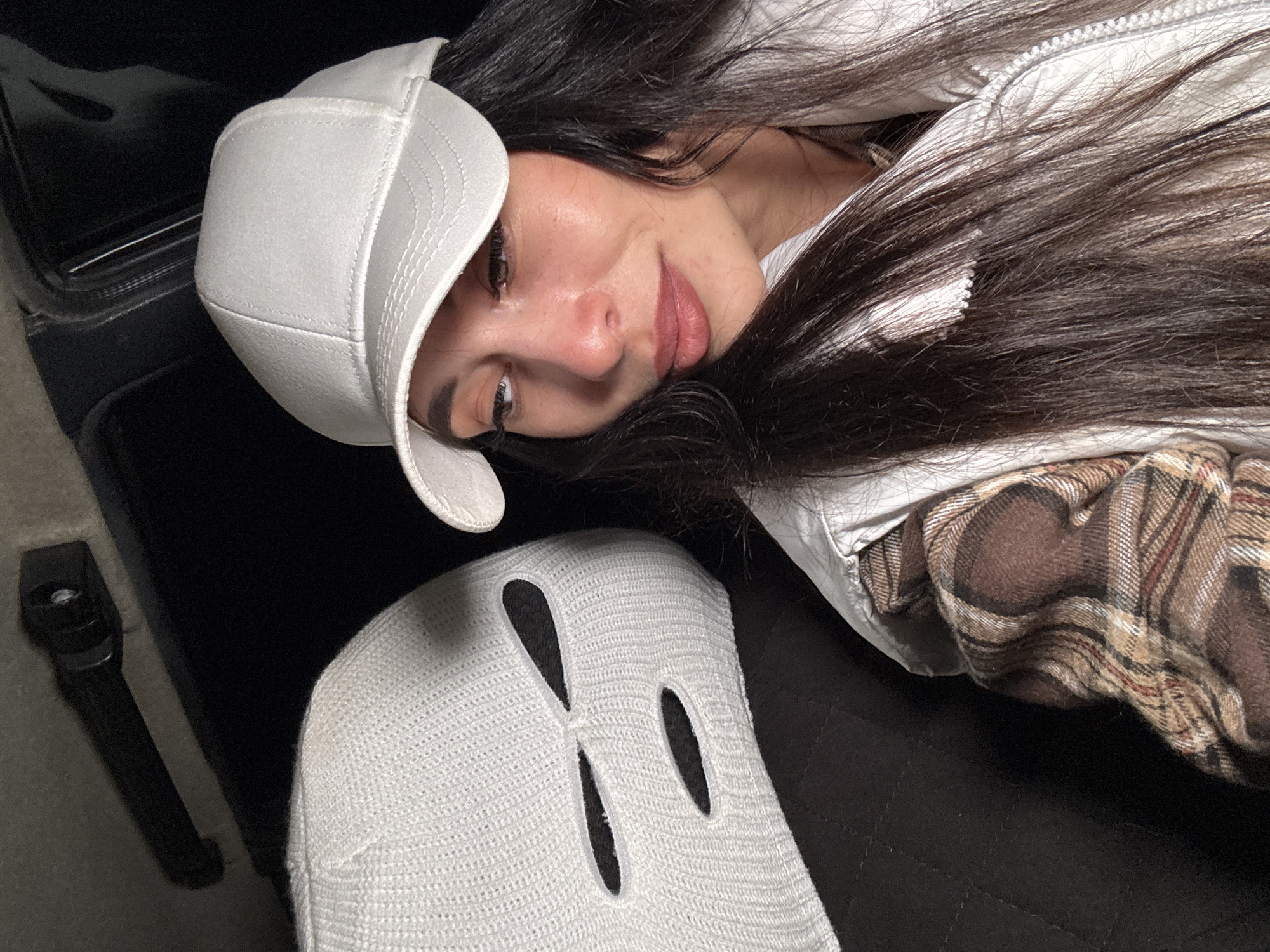 The image shows a woman with dark hair wearing a white baseball cap and a plaid shirt with a white vest. She is lying on a black surface, possibly a car seat, with another white hat with cut-out eye holes next to her. A black seatbelt buckle is visible to the left. There is no information in the image that indicates the location is Chaikovsky, Russia.