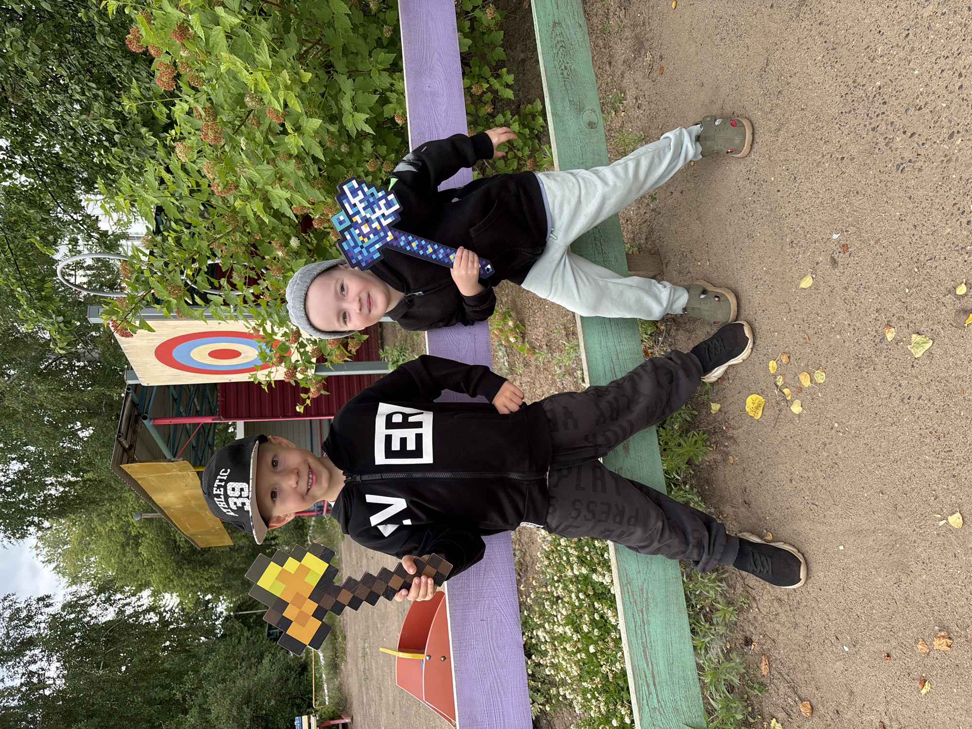 Two young boys, one wearing a black hoodie with "EVER" across the chest and a black baseball cap, and the other wearing a black hoodie and a grey beanie, stand in front of a wooden fence. The boy on the left holds a pixelated toy axe with a yellow and brown design. The boy on the right holds a similar pixelated toy axe, this one with a blue, white, and purple design. Behind them, a large target with concentric red, blue, and white circles is visible on a structure, along with lush green foliage and trees. The ground is covered in dirt and scattered yellow leaves. The scene appears to be outdoors, possibly in a park or recreational area. The presence of a target suggests an activity like archery or a carnival game. The overall impression is one of childhood play and outdoor recreation in a temperate climate, likely during the autumn season given the fallen leaves. The location is specified as the city of Chaikovsky, Russia, suggesting a common type of public space found in Russian cities.