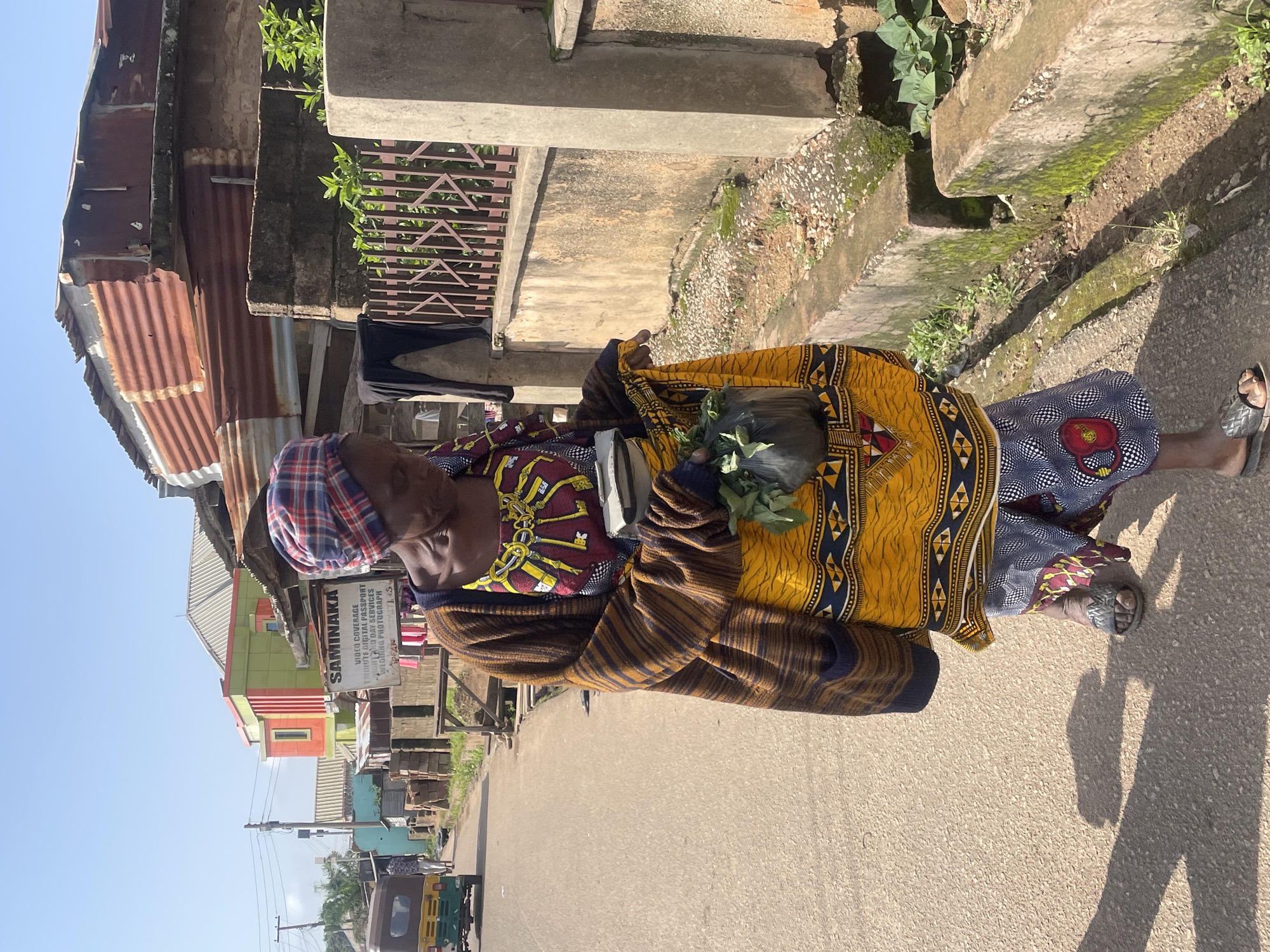 An elderly woman walks down a paved street in Jos, Nigeria, carrying a black plastic bag filled with green leaves. She is dressed in a vibrantly colored patterned skirt, a patterned top, and a striped cardigan. The street is lined with buildings, some with corrugated metal roofs, and a concrete drainage ditch. In the background, a colorful building with a modern design stands out, along with a sign for "SAMINAKA" which appears to be a business offering video coverage and photography services. A three-wheeled vehicle, commonly known as a Keke Napep, is also visible on the street. The sky is clear and blue, indicating a sunny day.