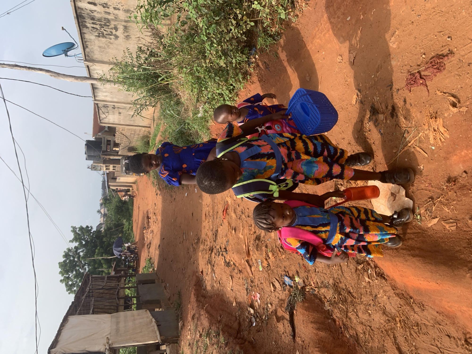 Three children and an adult stand on a reddish-brown, dirt path in Ukwu-Abwa, Nigeria. The adult wears a dark blue dress with orange flower shapes, while the children wear brightly colored orange and blue patterned dresses. The smallest child carries a blue plastic basket. One child holds a red plastic bottle and stands next to a white rock. A concrete wall with a blue satellite dish stands in the background on one side of the path, and a corrugated structure is on the other side. Vegetation grows along the wall. The sky is overcast.