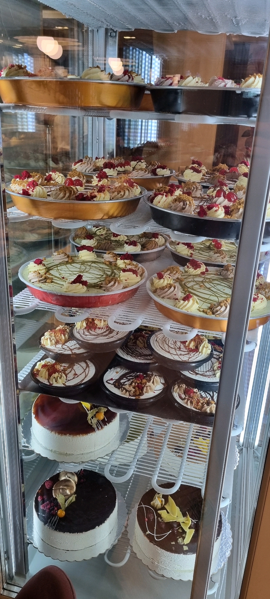 The image shows a refrigerated display case filled with various desserts. On the top shelf, there are several round pans containing what appear to be cheesecakes or tarts, topped with cream, berries, and crumble. The middle shelves are stocked with smaller, individual desserts in dark bowls, also adorned with cream, berries, and chocolate drizzle. The bottom shelf showcases three larger cakes. The cake on the left is round and white with a dark, glossy topping and a fruit garnish. The cake in the center is also round and white with a dark chocolate topping, decorated with berries, gold leaf, and a dried leaf. The cake on the right is similar, with a chocolate topping and a swirl of white icing, garnished with yellow decorative elements. The display case is well-lit, and the desserts appear to be freshly made and attractively presented. The overall scene suggests a bakery or dessert shop. The location is identified as Bashkia Durrës, Albania.