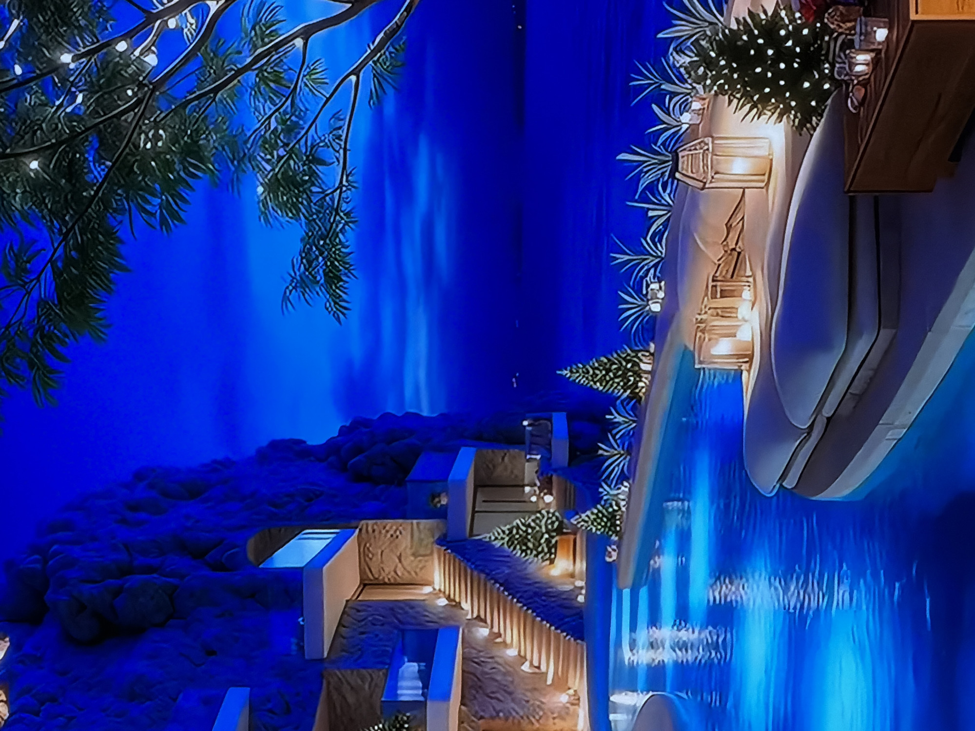 This media file captures a luxurious outdoor setting at night, characterized by a dominant deep blue color palette. The scene appears to be a multi-level resort or high-end leisure area, possibly in Sofiivska Borshchahivka, Ukraine.
In the upper left, a tree branch adorned with numerous small string lights extends into the frame. The background is a vast, dark blue expanse, suggesting either a night sky or a distant body of water.
On the right side, a pristine swimming pool or water feature is prominently featured, reflecting ambient blue light. Adjacent to the pool is a lounge or dining area with sleek white and possibly wooden furniture. This area is decorated with planters containing lush green foliage, resembling small festive trees, also illuminated with string lights. Elegant glass lanterns with glowing candles are arranged along a decorative railing or low wall, adding warm accents to the cool blue surroundings. A wooden structure, likely part of a table or bar, is visible in the upper right.
Towards the left and center, a series of modern, light-colored architectural structures, including pathways and stairs, ascend a natural, rocky hillside. These structures are subtly lit, guiding the eye upwards. More small, decorative trees with string lights are placed along these pathways. The rocky terrain itself is also bathed in blue light.
The absence of people suggests a tranquil and private atmosphere. The time of day is clearly night, emphasized by the dark backdrop and numerous artificial lights. The overall ambiance is serene, luxurious, and inviting, created by the careful integration of natural elements, modern design, and atmospheric lighting. No text is visible in the image.