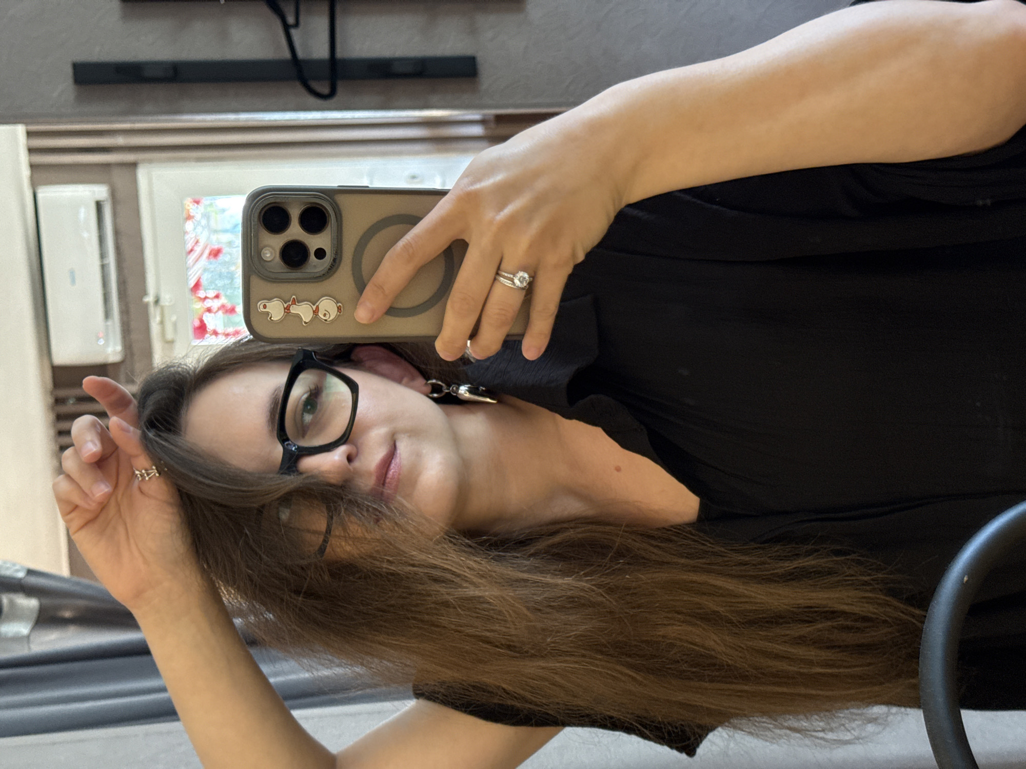 The image depicts a young woman taking a selfie in what appears to be a dressing room or bedroom. She has long, brown hair and is wearing black-rimmed glasses and a black shirt. Her left hand is raised, touching her hair, and she is wearing a ring on her left ring finger. Her right hand is holding a smartphone, which has a phone case with three small duck stickers and a circular pattern. She is also wearing a silver earring on her left ear. In the background, there is a mirror reflecting an air conditioning unit and a window with curtains. There is also a dark hanger in the reflection on the right. The scene does not provide any specific details or context that would suggest a location in Chaikovsky, Russia.