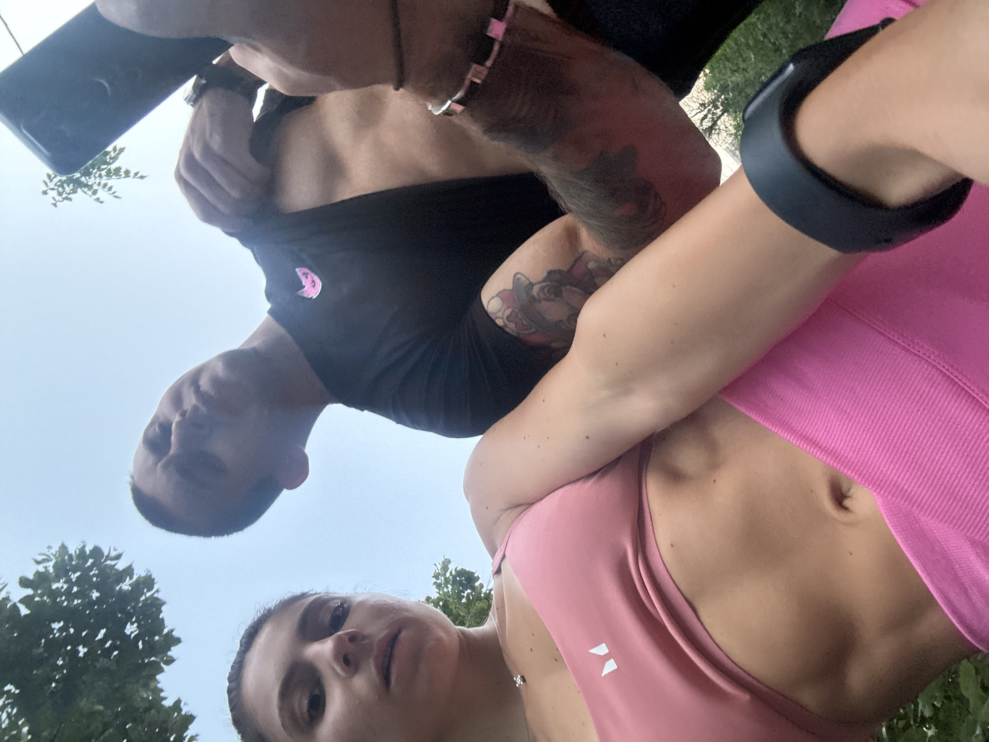 An outdoor, low-angle, waist-up selfie features two individuals. The person on the left, facing the camera, is a woman with dark hair and a pink sports bra. Her abdominal muscles are visible. The person on the right is a man with a black t-shirt and visible tattoos on his arm. He is holding a smartphone, presumably to take the photo. They are both outdoors, with greenery and a light blue sky visible in the background. The lighting suggests it is daytime. There is no specific information in the image to place the location in Varna, Bulgaria.