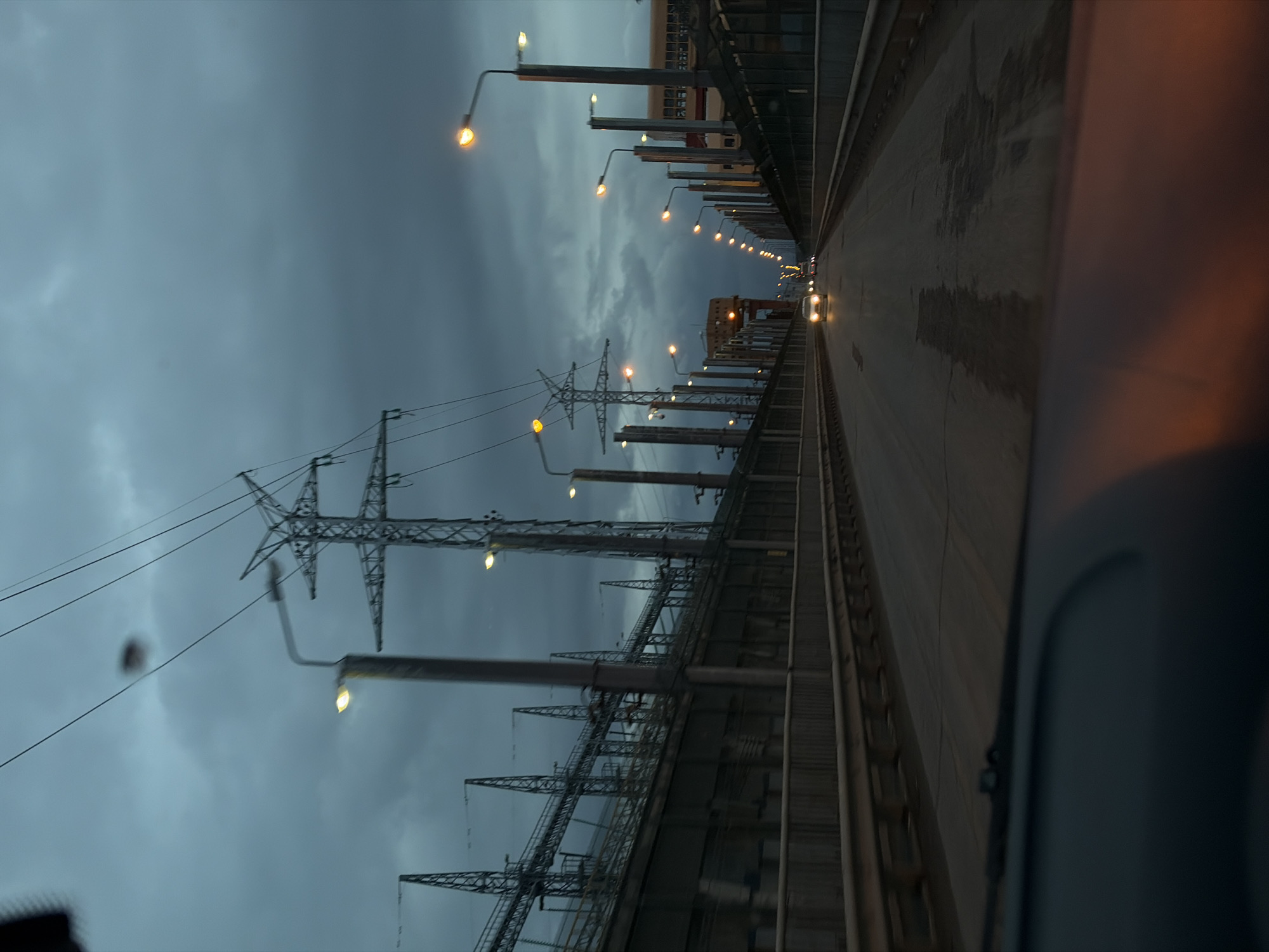 The image, taken from within a moving vehicle, depicts a wide industrial landscape at dusk or early night in Chaikovsky, Russia. The sky is heavily overcast with dark grey-blue clouds, creating a somber atmosphere.
Dominating the left side of the frame are several towering electricity pylons and a complex network of power lines, indicating a significant power generation or transmission facility. These structures stand starkly against the gloomy sky.
On the right, a long, straight road or bridge extends into the distance, flanked by a continuous row of streetlights. These lights emit a warm, yellow-orange glow, illuminating the road surface and adjacent industrial structures. The light from the streetlights creates a stark contrast with the dark sky and industrial forms. In the far distance, the bright headlights of another vehicle are visible, traveling along the same path.
The perspective from the vehicle's interior, with a blurred dark shape on the far right suggesting a window frame or dashboard, adds to the sense of movement. The overall scene conveys the scale and functionality of a major industrial area, likely a hydroelectric power plant or dam, which aligns with Chaikovsky's location near the Votkinsk Hydroelectric Power Plant. There is no visible text in the image.