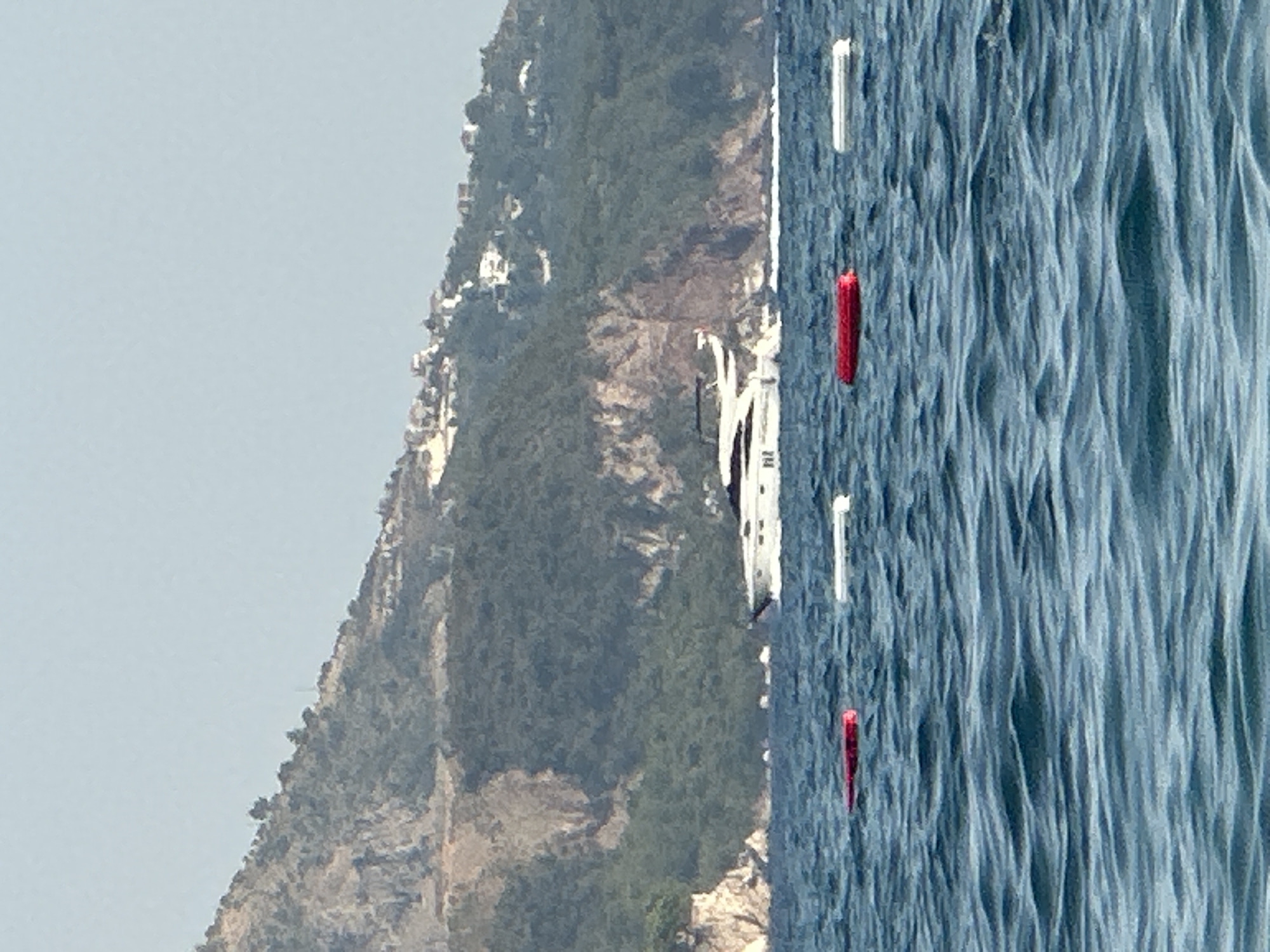 A large white yacht is cruising on the wavy blue sea in front of a verdant, mountainous coastline. Several white and red buoys are floating in the foreground of the image, marking a section of the water. The coastline is dotted with white buildings, suggesting a populated area. The sky is a clear, pale blue. The scene appears to be a summer day in Boreti, Montenegro.