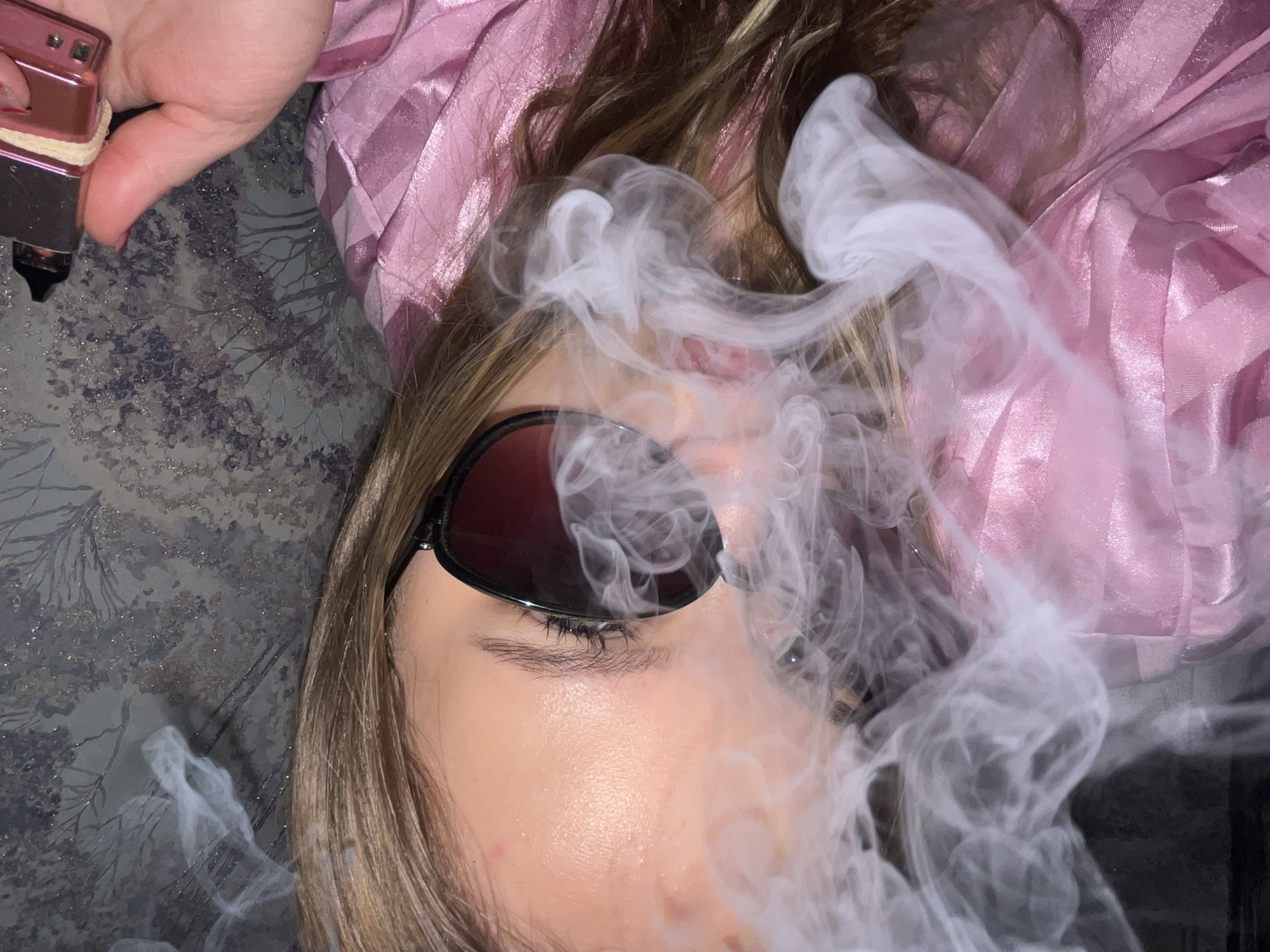 A person with light brown hair is lying down, wearing sunglasses and a pink striped garment. Smoke is present in the foreground, obscuring parts of the person's face. A pink and silver device is visible in the upper left corner. The background features a gray and white patterned surface. The location is not discernible from the image.