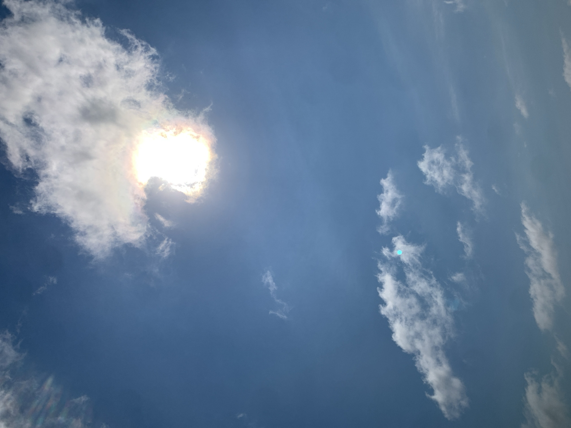 The image displays a bright, sunlit sky with scattered clouds. The sun is partially obscured by a cloud, creating a halo effect with iridescent colors. The sky is a clear, deep blue. In the lower portion of the image, there are more clouds, some appearing wispy and others denser. A small, bright blue circular anomaly is visible within one of the lower clouds. The scene is an outdoor, atmospheric view without any discernible human presence, structures, or specific landmarks that would indicate a location within Khazar Raion, Azerbaijan.