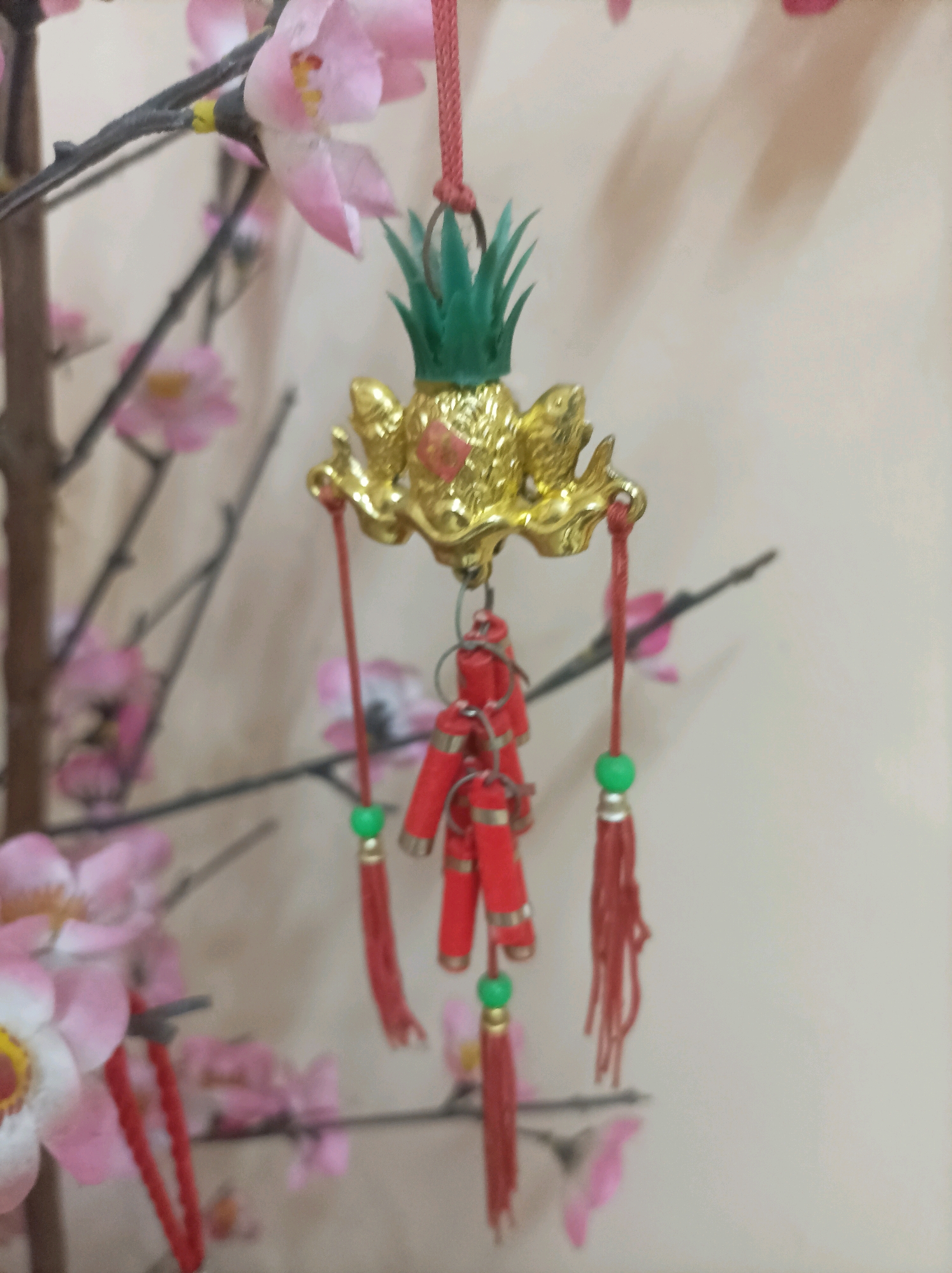 A decorative ornament hangs from a branch of a faux floral arrangement. The ornament features a golden, ornate top section with what appears to be a frog or dragon-like creature, and a green pineapple-shaped flourish. Dangling from this top piece are several strings of small, red, firecracker-like decorations, each adorned with small green beads and red tassels. The background is a light-colored, textured wall, with the blurred pink blossoms of the floral arrangement providing context. The image quality is moderate, with a shallow depth of field that blurs the background elements and highlights the central ornament. The setting appears to be indoors, possibly a home or establishment in Bekasi, Indonesia, given the context provided.