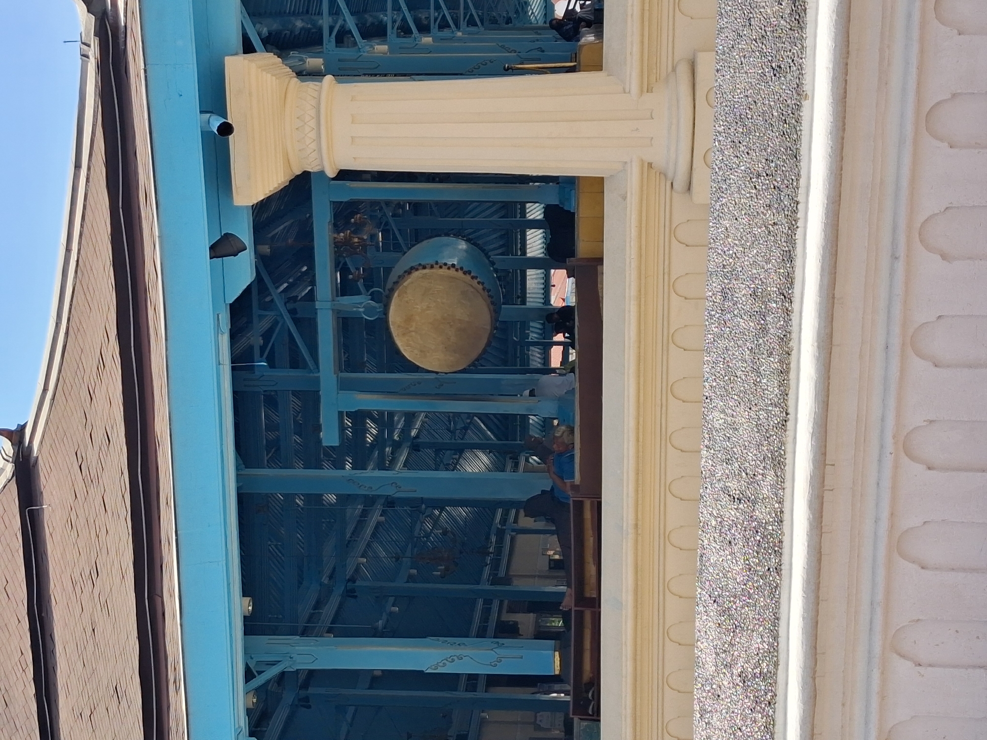 The image shows a large, light-colored, cylindrical drum suspended from a blue wooden frame under the roof of a building. The drum is ornately decorated with small studs along its circumference. The building has a brown tiled roof and a light blue exterior structure supported by cream-colored fluted columns. In the background, several people are visible, some sitting and others lying down, inside the open-air structure. The ground in front of the structure is covered with small grey pebbles. This scene is likely within the grounds of the Surakarta Sunanate Palace (also known as Keraton Surakarta Hadiningrat) in Surakarta, Indonesia, where large drums, known as "gamelan" drums, are often found as part of cultural and religious ceremonies. The overall atmosphere suggests a peaceful and possibly sacred setting.
