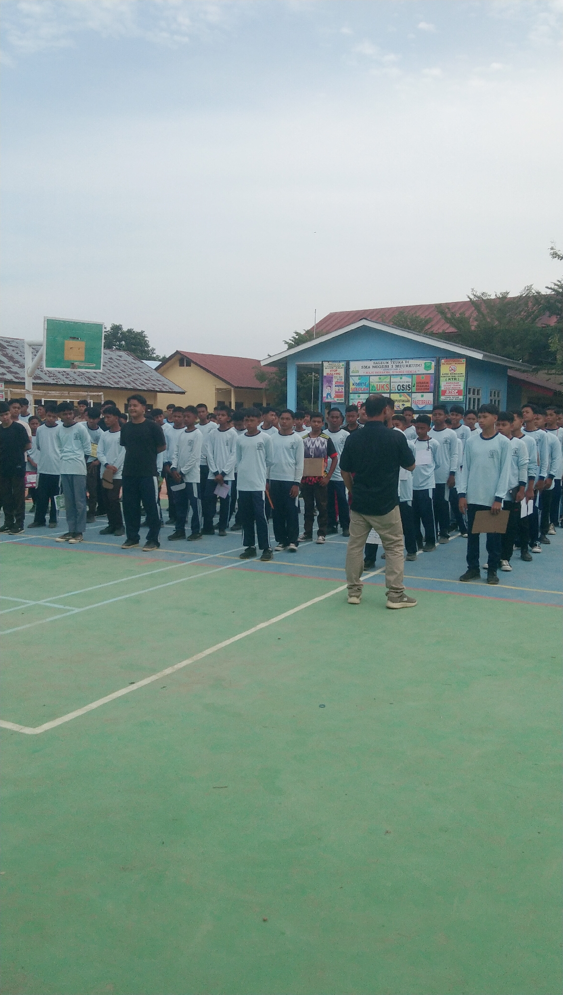 The image depicts a scene at SMA Negeri 1 Meureudu, located in Rhieng Blang, Indonesia. A group of young males, likely students, are assembled on an outdoor court. Most are wearing light blue long-sleeved shirts and dark pants. One individual, standing slightly apart and facing the group, is wearing a dark shirt and light-colored pants, appearing to address or supervise them. Behind the assembled students, a light blue building with a red roof is visible, bearing signage that includes "SMA NEGERI 1 MEUREUDU" and "OSIS," indicating it is a school building. A basketball hoop with a green backboard is also present on the left side of the frame. The court itself is green with white markings, suggesting it is used for sports activities. The sky is overcast with some visible clouds.