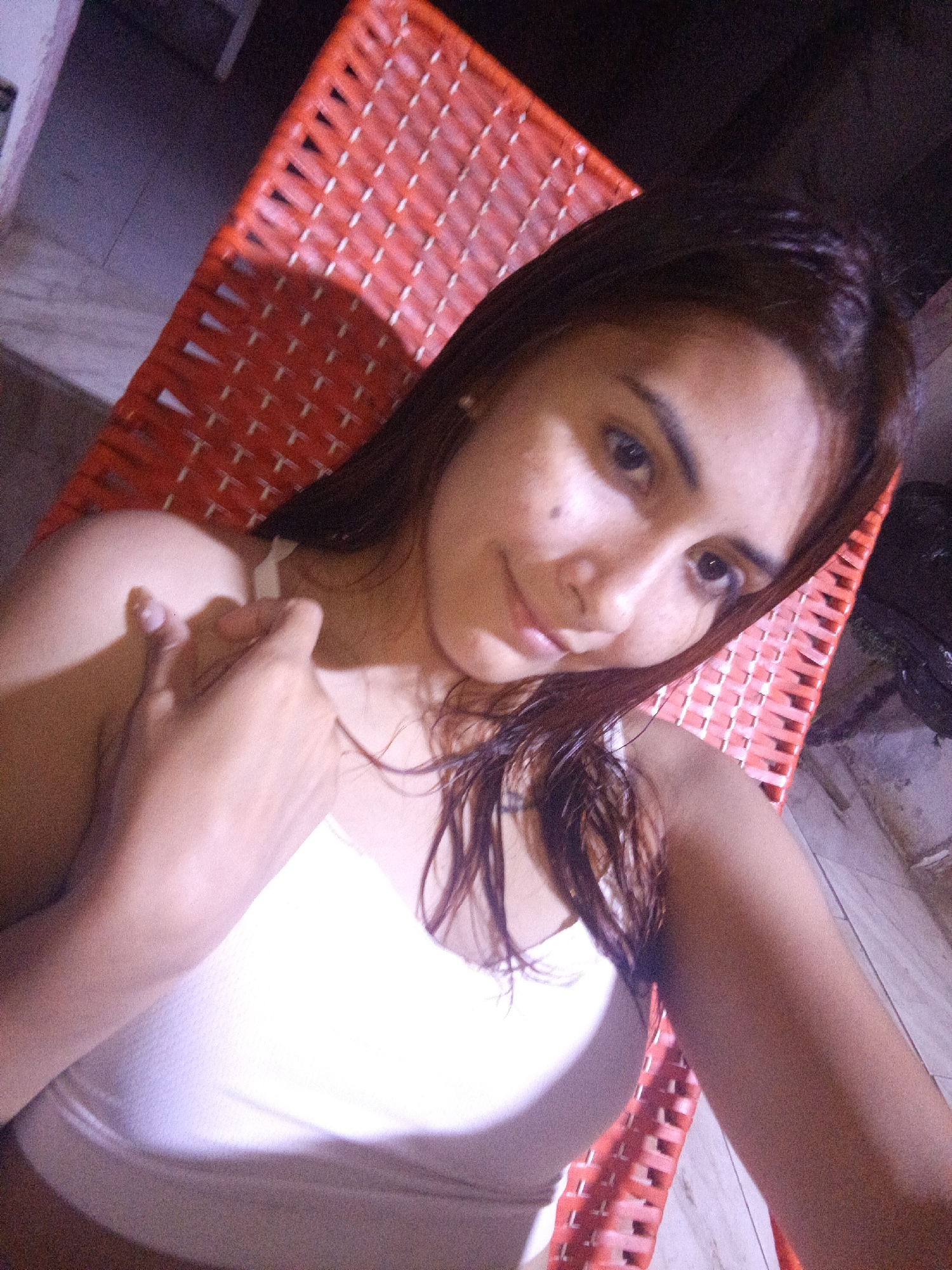 A young woman is reclining in a red, woven chair. She is wearing a white tank top and has shoulder-length brown hair, some of which is damp and clings to her face. Her hand is raised near her shoulder. The setting appears to be an outdoor space, possibly a patio or terrace, with tiled flooring. The location is possibly in or near Juan Griego, Venezuela.