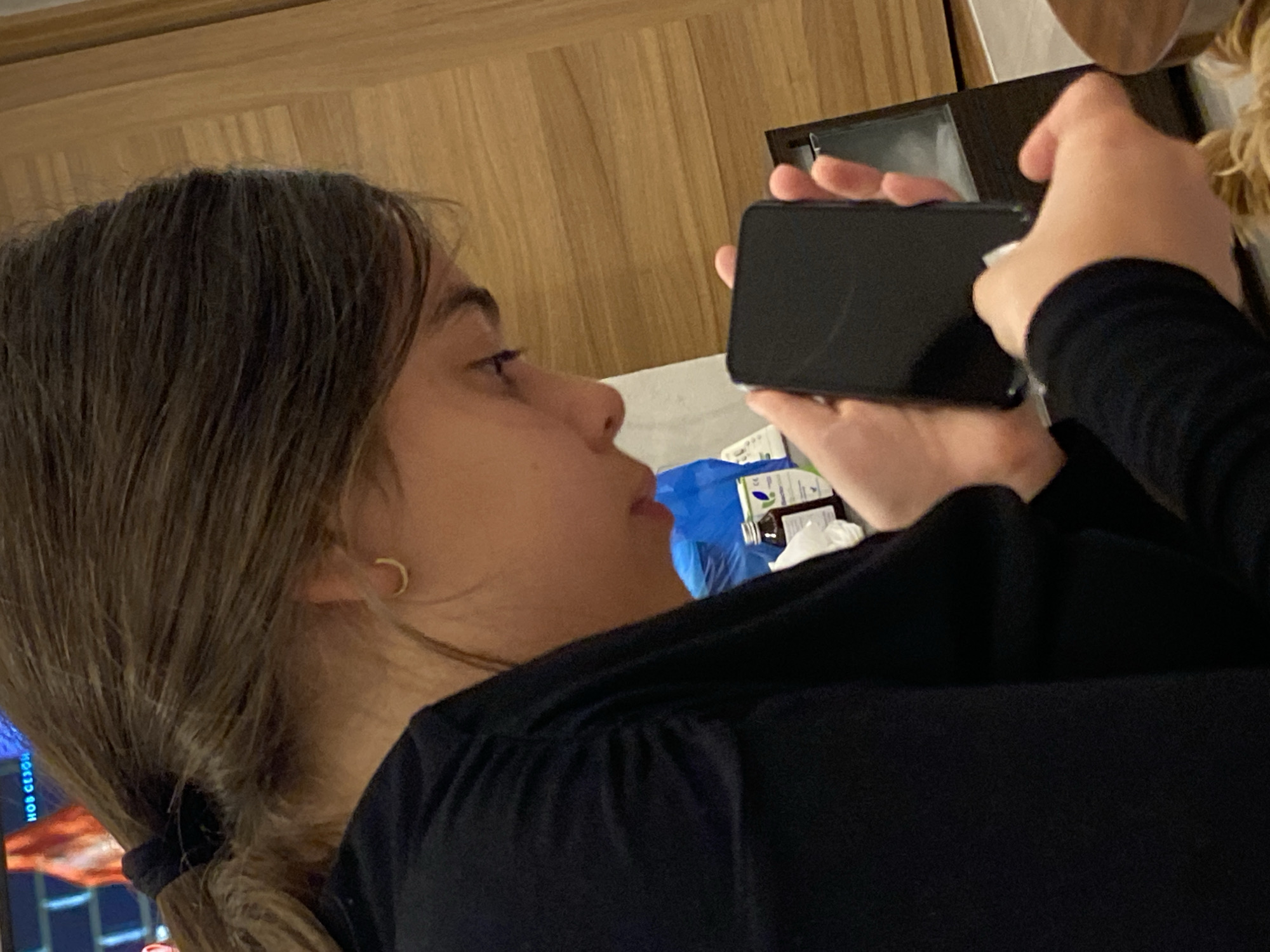 A person with long, dark brown hair, pulled back into a ponytail, is holding a black smartphone and appears to be cleaning the screen with a white wipe or cloth. The individual is wearing a black long-sleeved shirt. The background includes a wooden wall, a blue bag, and what seems to be a box of medication. The location of the image is likely inside a building, given the interior setting. There is no information indicating that the image was taken in Dragovishtitsa, Bulgaria.