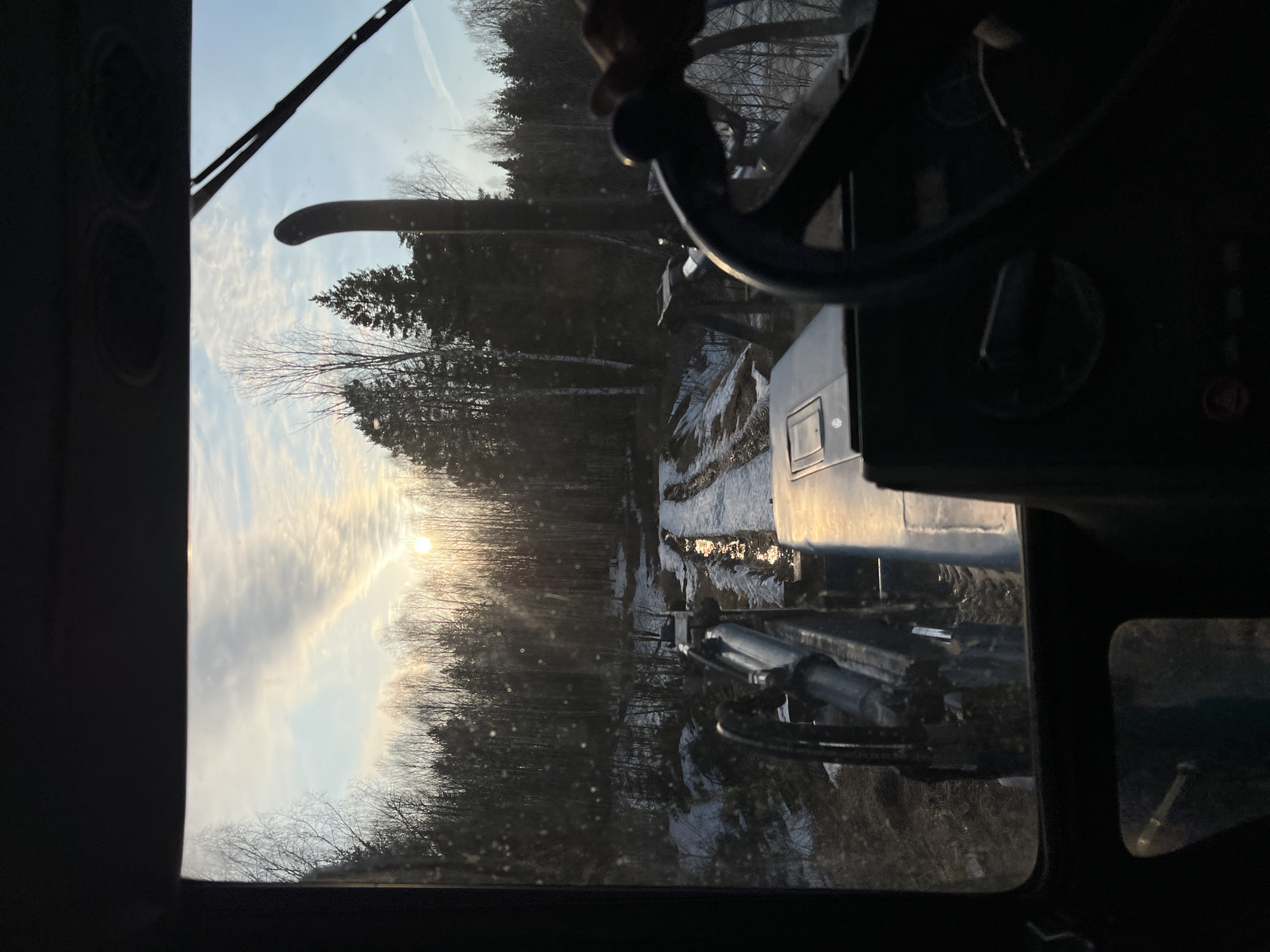 A view from inside a vehicle, likely a tractor or heavy machinery, facing a wooded area. The sun is visible through the trees. Patches of snow are visible on the ground. The location is uncertain but the caption suggests that the photo was taken in Zhigalki, Russia.