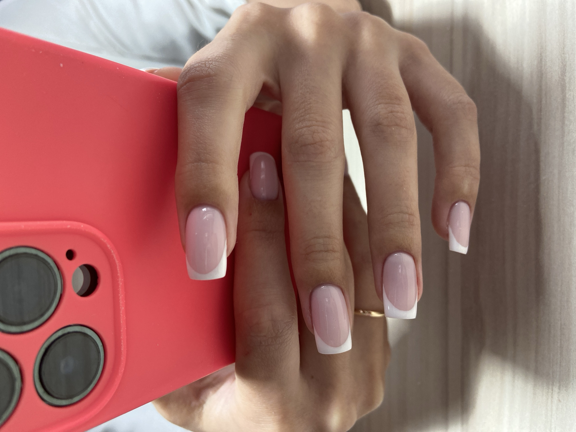 A close-up photograph shows a hand holding a red iPhone, with the fingers extended. The fingernails have a classic French manicure with a pale pink base and a white tip. A gold ring is visible on the ring finger of the hand. The iPhone has three camera lenses and a smaller hole presumably for a microphone or flash. The background is a light, textured surface resembling wood. The photo does not contain any location in the city of Budva, Montenegro.