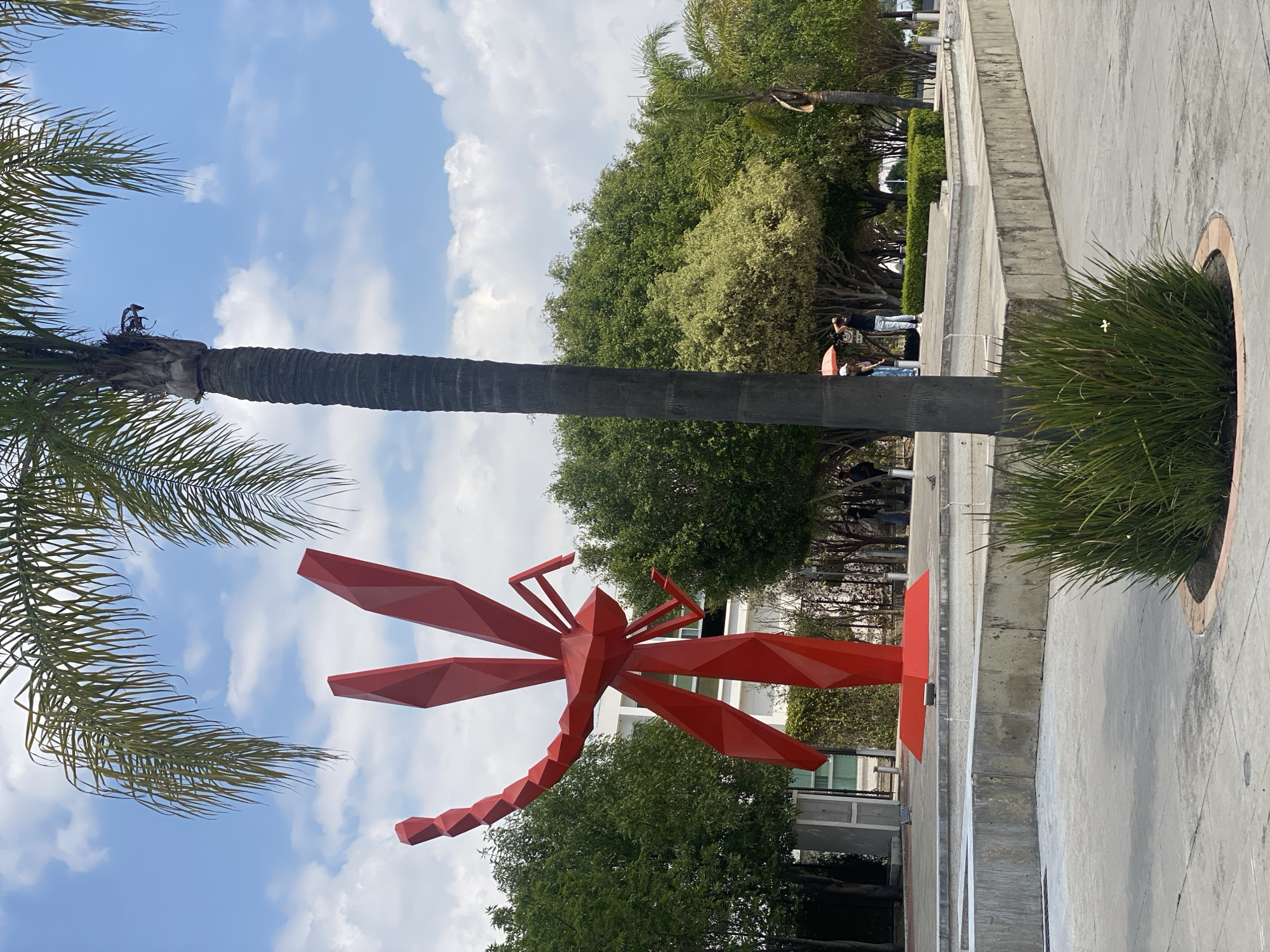Observed in an outdoor setting in an unidentified urban location, a large red geometric dragonfly sculpture stands prominently next to a palm tree. Behind the sculpture are green trees and what appear to be buildings. To the side, there's a raised concrete platform with plants in a circular planter. In the background, near the tree line, are two people standing. The sky is blue with scattered clouds.