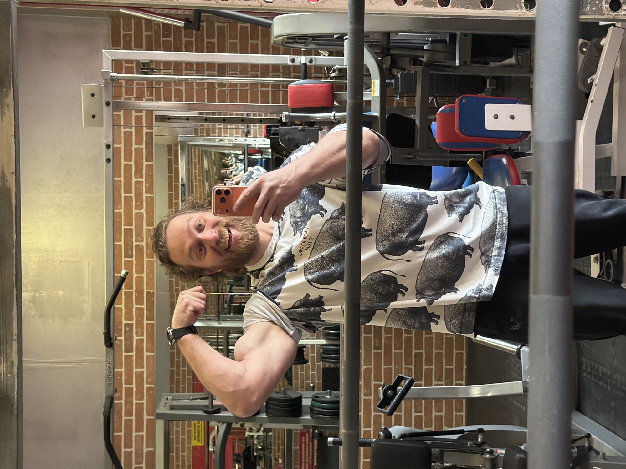 The image shows a man taking a mirror selfie in a gym located in Saint Petersburg, Russia. He has curly brown hair and a beard, and is smiling broadly at his orange-brown smartphone, which he holds in his left hand to capture the photo. He wears a white t-shirt patterned with black illustrations of pigs or boars, and dark pants. His right arm is bent, flexing his bicep, and a dark-strapped watch is visible on his left wrist.
The gym interior is reflected behind him, featuring brick-patterned walls and various exercise equipment, including weight stacks, bars, and machines with padded elements in red, blue, and white. The man's cheerful expression indicates a positive and confident mood. On his shirt, partially visible, mirrored text resembles "MAI HOHIC," likely an inverted slogan.