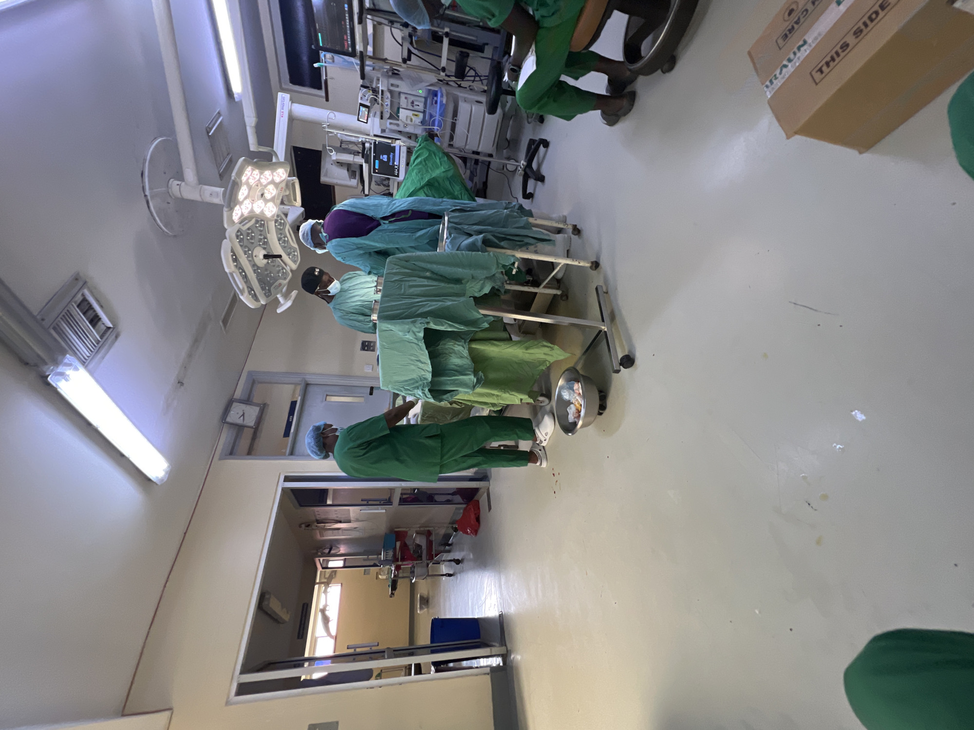 The image depicts a surgical operating room, possibly located in Nairobi, Kenya. The room is brightly lit with overhead surgical lights. Several individuals dressed in green surgical scrubs are present, some wearing surgical masks and caps. They appear to be attending to a patient on an operating table covered with green surgical drapes. Medical equipment and monitors are visible in the background. A cardboard box labeled "BRAUN" and "THIS SIDE" is also present in the room. A doorway leads to another area with additional medical equipment. The environment appears sterile and organized.