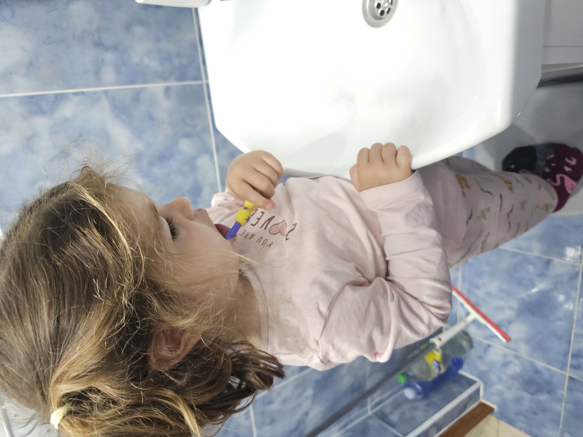 A young girl with light brown hair tied in a ponytail is brushing her teeth in a bathroom. She is wearing a pink long-sleeved shirt with a heart and text on it, and patterned pajamas. A yellow and purple toothbrush is in her mouth. She is standing next to a white sink. The bathroom walls are tiled with blue and white squares. The floor is also tiled in blue. In the background, a blue bottle and a white squeegee are visible. The image is a close-up, focusing on the girl and the sink.