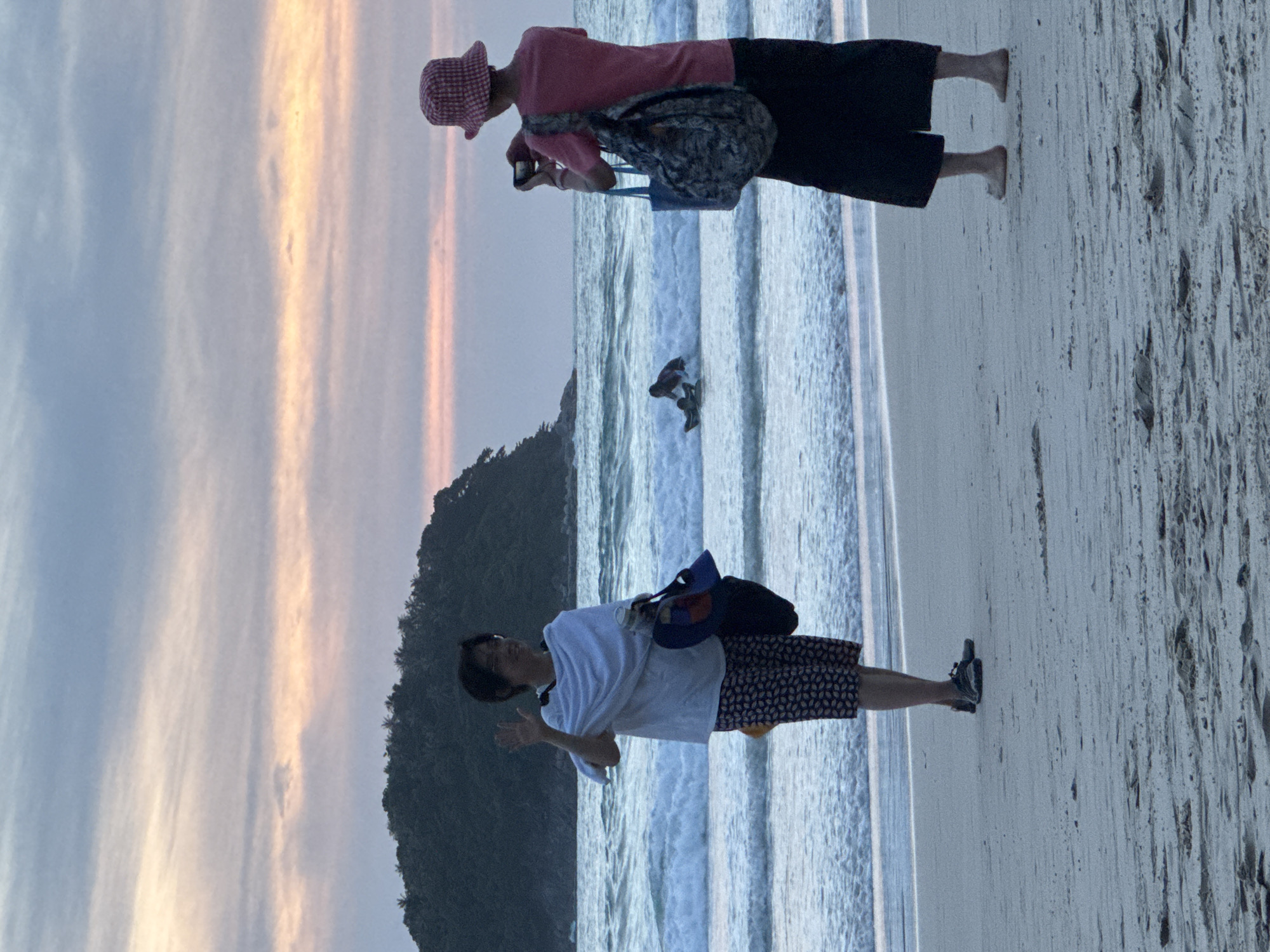 Two individuals are on Karon Beach in Karon, Thailand, at dusk. The sky is filled with soft pink and orange clouds. In the foreground to the left, a person wearing glasses, a white draped cloth over their shoulders, and patterned shorts waves at the camera. They are holding a bag and a water bottle. To the right, another person wearing a pink checkered hat and a pink t-shirt, and black pants, is taking a photo with a phone. They have a patterned bag slung over their shoulder. In the background, the ocean waves are crashing, and a person is visible on a surfboard. A lush, green island is in the distance.