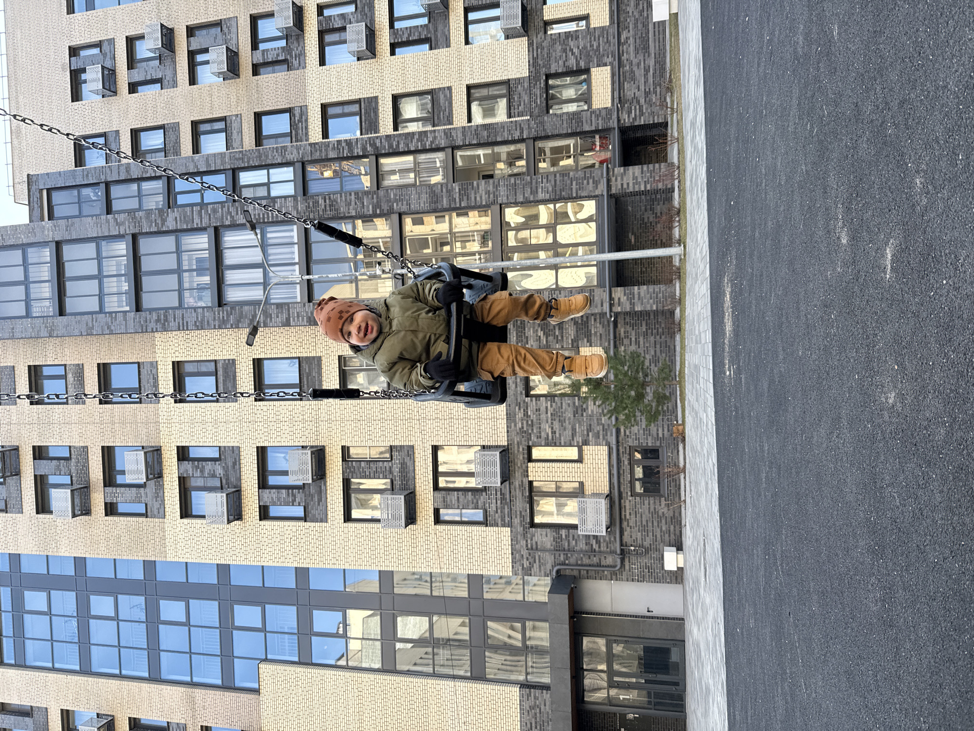 A child is swinging on a swing set in front of an apartment building in Rusanovo, Russia. The child is wearing a brown hat, green jacket, and brown pants. The apartment building has a mix of tan and gray brick. Some windows have open metal ventilation panels. The ground surface appears to be asphalt and grey tiles.