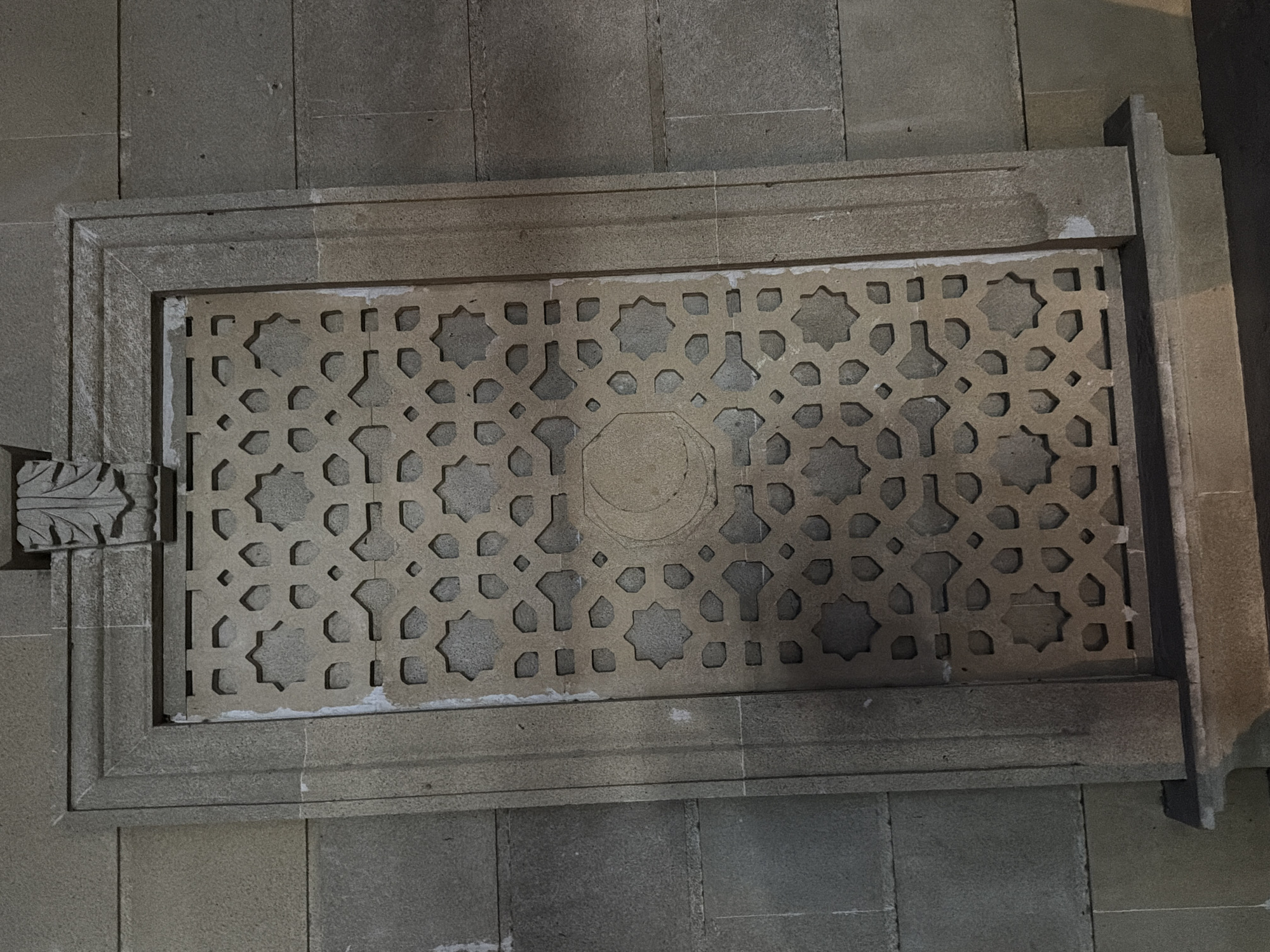 The image shows a close-up of a stone facade with a decorative geometric pattern. The pattern is carved into a rectangular panel that is set within a larger stone frame. The panel features interlocking star shapes and other geometric forms, with a central circular motif containing a crescent moon. The stone is a light brown or beige color, and the surrounding wall is constructed of larger, rougher stone blocks. The lighting suggests it is either dusk or an interior setting with artificial light. There are no people or animals visible in the image. The image is taken from a straight-on perspective, with a slight downward angle. The overall impression is one of architectural detail and possibly historical significance. The location of Salyan, Azerbaijan is not visually evident from the image itself, but if this is indeed a landmark in that city, it would be a component of its architectural heritage.