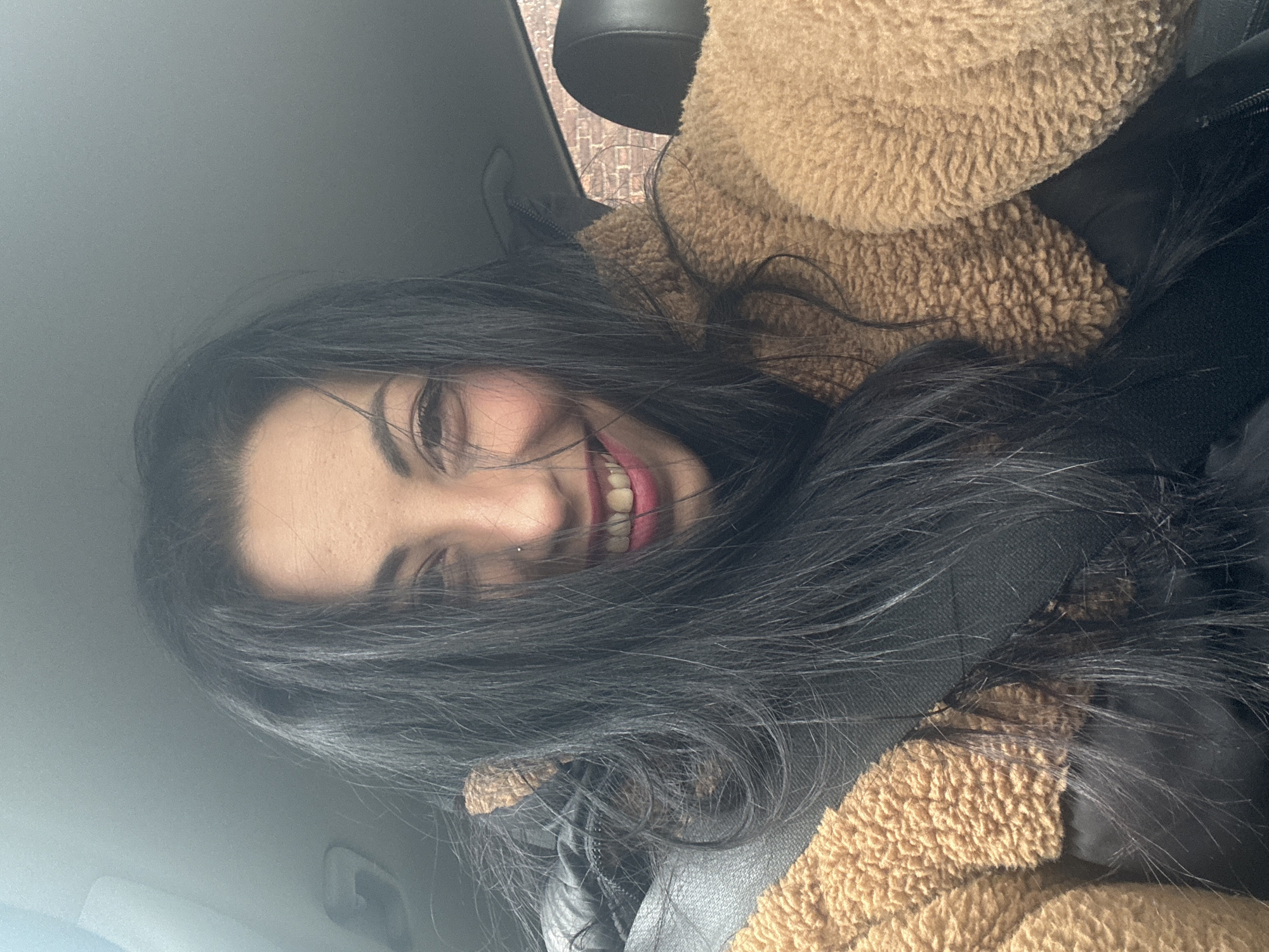 A woman with long, dark hair smiles towards the camera, partially obscured by her hair. She appears to be wearing a brown, textured jacket and a black top or scarf. The setting appears to be inside a car, indicated by the car interior elements visible. The location within Warrington, United Kingdom, is not discernible from the image.