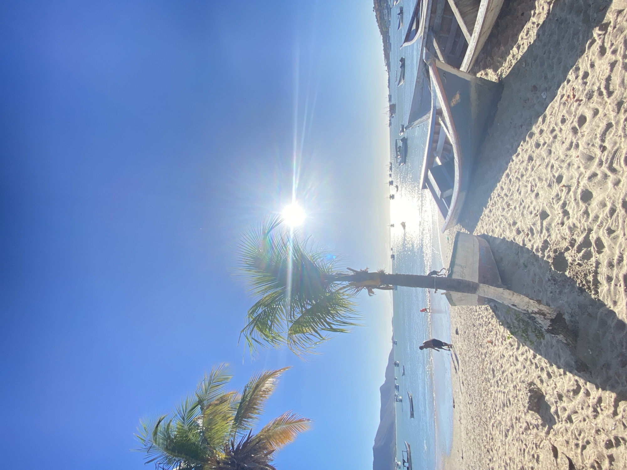 The image depicts a beach scene in Juan Griego, Venezuela, on a sunny day. In the foreground, two palm trees are visible against a bright blue sky with the sun shining brightly, creating a lens flare effect. A partially obscured figure of a person is walking on the sandy beach. A line of fishing boats are pulled up on the beach. Further out in the water, more boats are anchored. A landmass, possibly a mountain, is visible in the background. The photograph is taken from a low angle, giving prominence to the sky and the palm trees. The colors are vibrant and the lighting is strong.