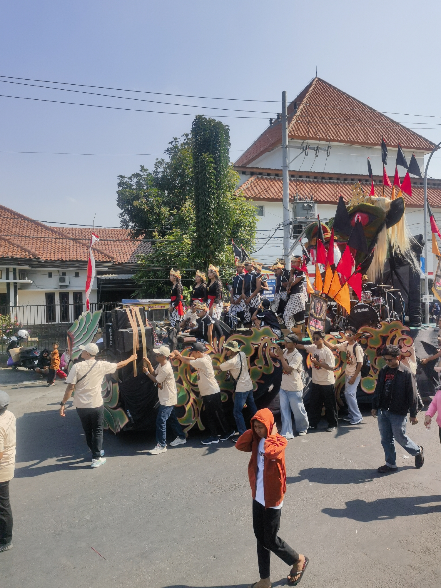 This image captures a vibrant street scene in Blora, Indonesia, likely during a cultural event or parade. In the foreground, several individuals are walking on an asphalt road, casting long shadows under a clear blue sky. One person in an orange hooded jacket and black pants is prominently featured, appearing to adjust their headphones. To the right, a person in a black jacket and jeans walks past.
The central focus of the image is a large, elaborately decorated float or stage. It is adorned with what appears to be a dragon or mythical creature head at the front, complete with large eyes and a flowing mane. Colorful flags, predominantly red and orange, are attached to poles on the float, adding to the festive atmosphere. Several people are visible on and around the float, some dressed in traditional attire with ornate crowns, while others are engaged in operating or observing the structure. A drum set with a "YAMAHA" logo is also visible on the float.
Behind the float, buildings with red-tiled roofs are visible, suggesting a developed urban area. Green trees are scattered throughout the background, interspersed with utility poles and power lines. The overall impression is one of a lively, public gathering taking place on a sunny day in Blora.