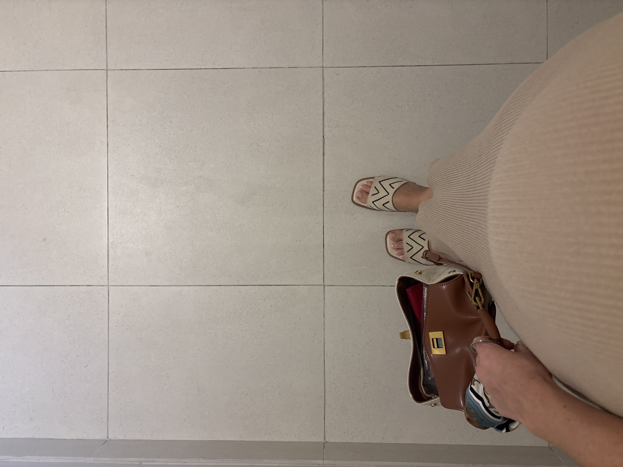 A top-down perspective captures a portion of a person standing on a light-colored, tiled floor. The person is wearing a long, light-beige, ribbed dress and sandals with a black and white zigzag pattern. A brown handbag with a gold clasp and a colorful scarf attached is visible near the person's hand. The scene is neutral, with the tiled floor dominating the left side of the frame. The location in Choeng Thale, Thailand is not discernible from the image.