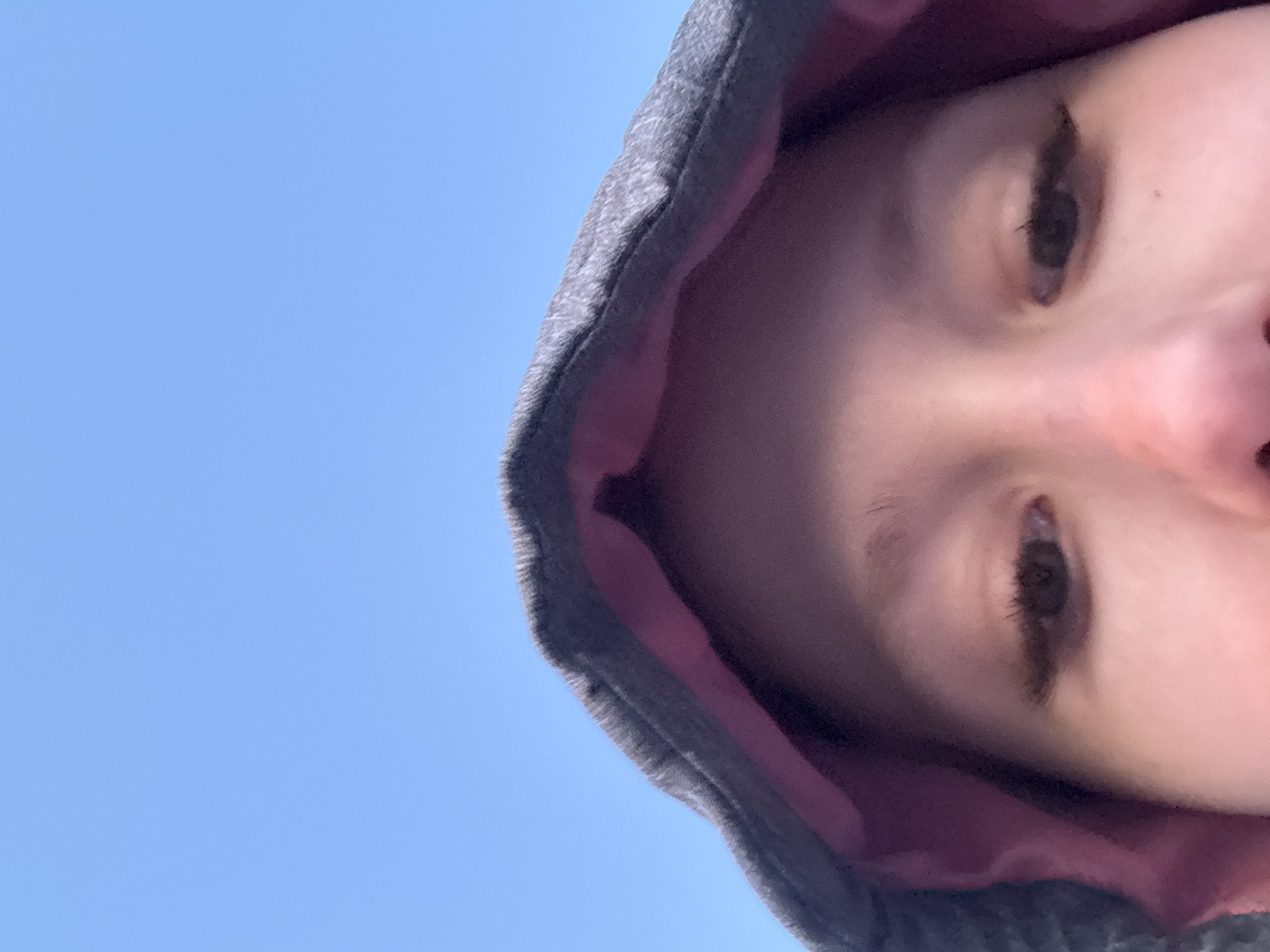 The image shows a close-up view of a person's face from a low angle, framed by the hood of a gray hoodie with a pink lining. The person's eyes are visible, gazing downwards. The background is a clear, light blue sky. There is no contextual information in the image to establish a location in Berezniki, Russia. The image lacks specific details of place, actions, or interactions other than a person wearing a hoodie.