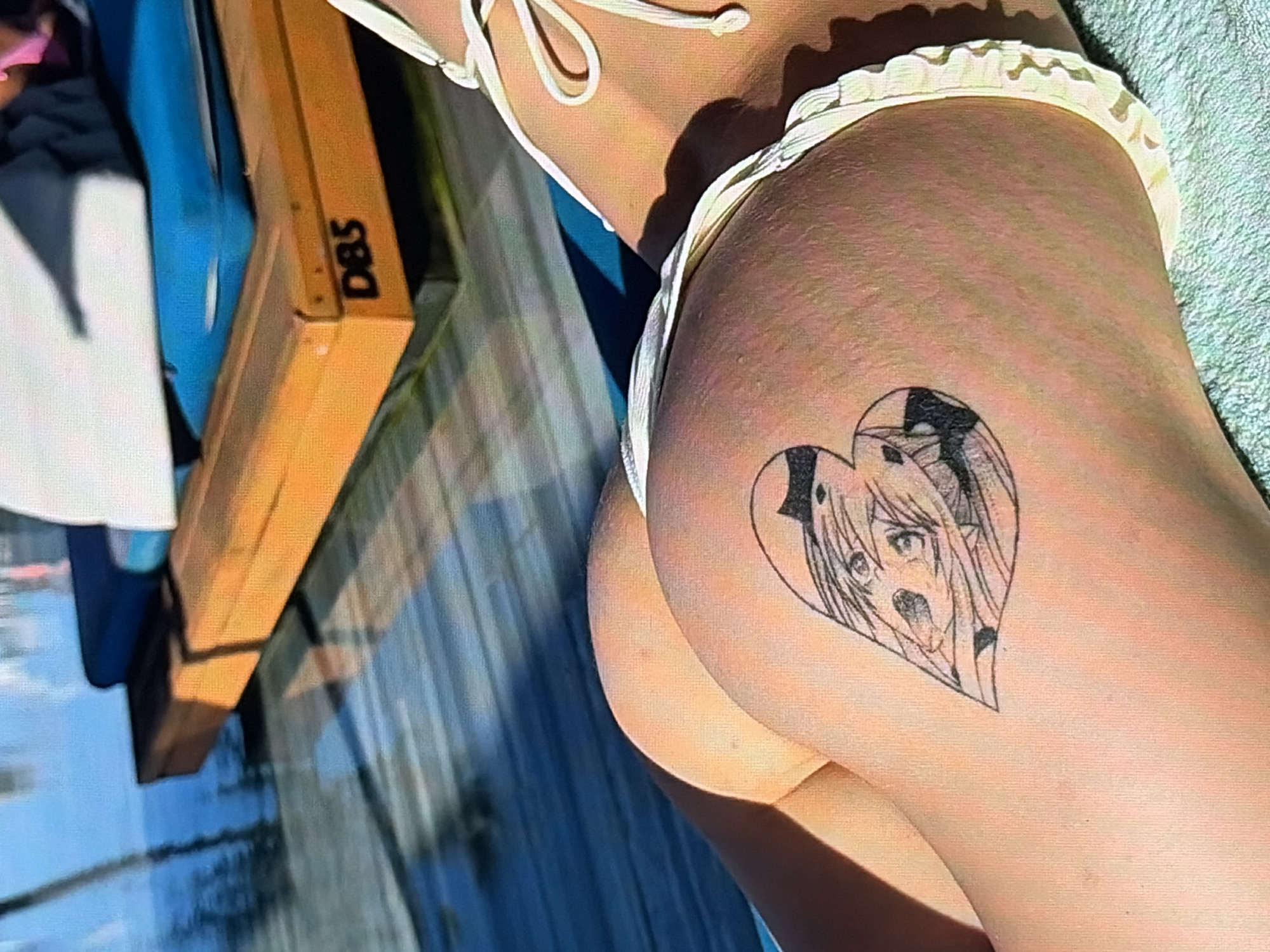 The image shows a close-up view of a person's buttocks with a heart-shaped tattoo. The tattoo depicts two anime-style characters. The person is wearing a white bikini bottom with ruffled edges. In the background, a wooden lounge chair with blue cushions is visible, situated on a wooden deck next to a body of water, possibly a swimming pool. The lighting suggests it is daytime. The scene appears to be an outdoor setting, likely a resort or a poolside area.