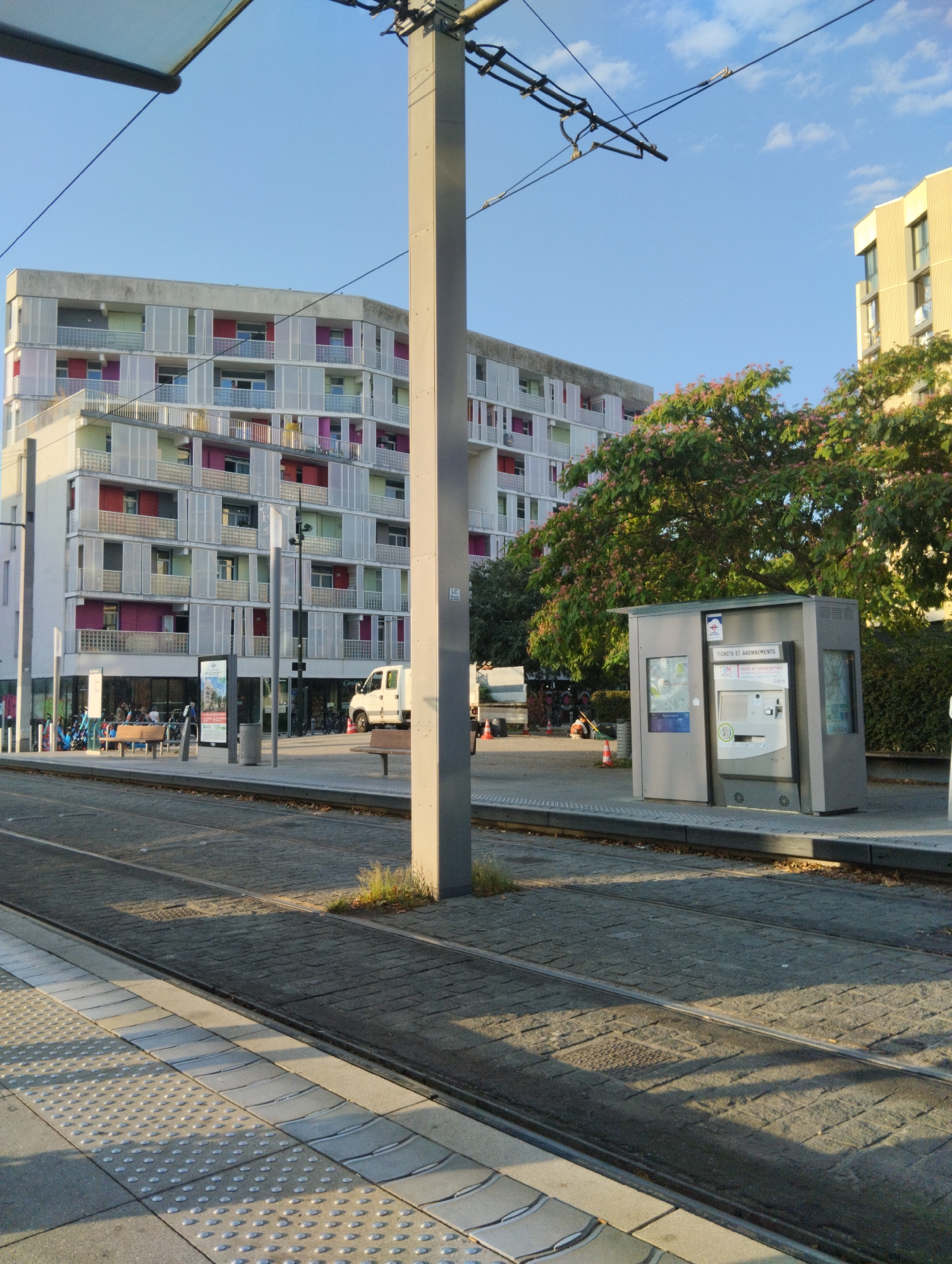 The image depicts a tram stop in Bègles, France, on a clear day with blue sky and scattered clouds. In the foreground, a tram track runs horizontally across the scene, bordered by paved pedestrian areas. To the left, a section of a tram platform is visible with textured paving.
Dominating the background is a modern, multi-story building with a distinctive facade featuring alternating light-colored panels and colorful balconies in red and pink. Adjacent to this building, on the right side of the image, is a ticket vending machine. A white van is parked near the building's entrance.
Several individuals are visible, some seated on benches near the building, others standing or walking. A large tree with pinkish flowers is in bloom between the modern building and the ticket machine. Overhead, tram wires and a support pole are prominent. The overall scene suggests a functional urban public transport area bathed in sunlight.