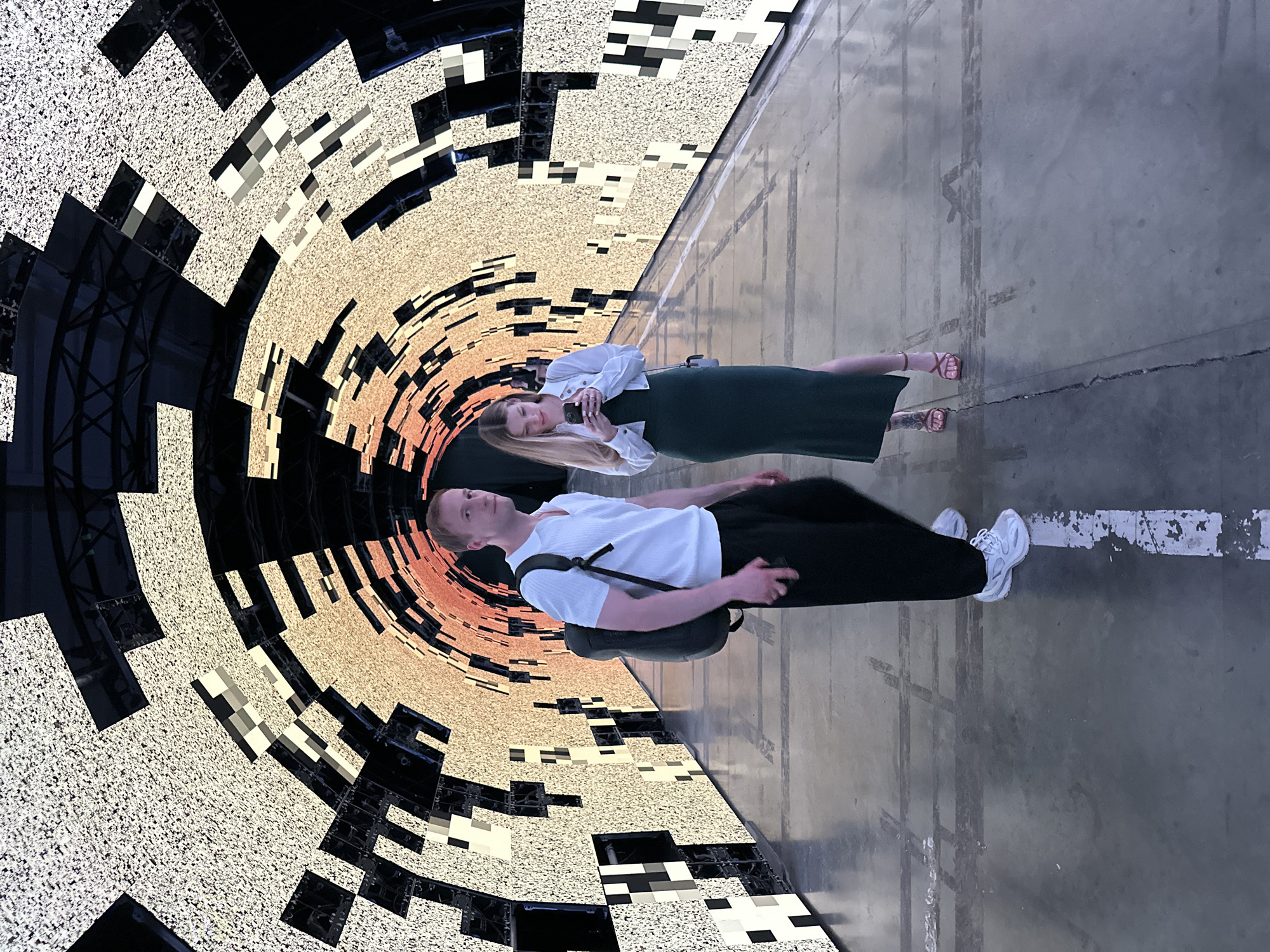 This media file captures a man and a woman standing in an urban environment. The image is oriented such that the subjects appear to be standing on a mirrored surface, with their reflections visible below them. The background features a striking, abstract geometric design, reminiscent of a tunnel or a circular vortex, composed of textured light and dark squares and rectangles in shades of beige, black, and orange.
The woman, on the left side of the frame, is wearing a white collared shirt, a dark green dress, and pink sandals. She is holding a smartphone and appears to be taking a picture or recording a video. Her hair is blonde and falls past her shoulders.
The man, on the right side of the frame, is dressed in a white t-shirt, black pants, and white sneakers. He has a black backpack slung over his shoulder and is holding a smartphone in his hand. He is looking to his left, slightly away from the camera.
The setting appears to be indoors, possibly an art installation or a public space with a visually dynamic display. The floor beneath them is a smooth, gray concrete surface marked with faint lines and scuff marks, suggesting a utilitarian or industrial space. The lighting is bright and diffused, creating reflections that enhance the surreal quality of the scene. The overall mood is modern and perhaps a little disorienting due to the visual effects and reflections. There is no visible text in the image. The context provided indicates this scene is in Saint Petersburg, Russia.