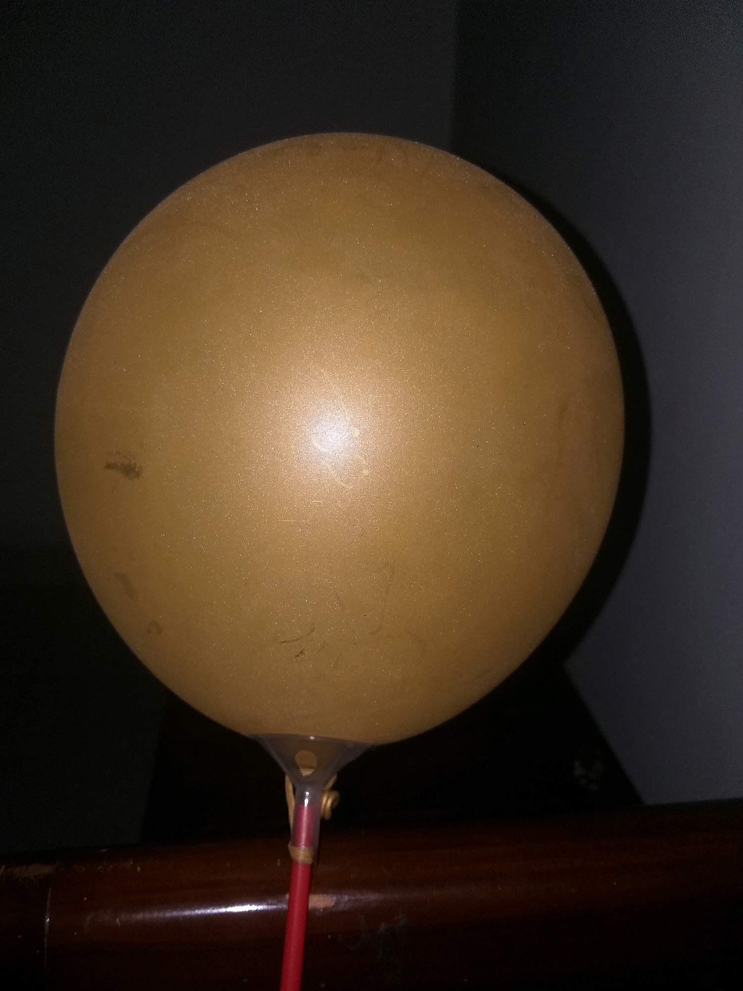 A close-up, slightly angled shot shows a tan balloon against a dark, out-of-focus background. The balloon is inflated and rounded, with a subtle sheen and some minor scuff marks visible on its surface. At the base of the balloon, a clear plastic holder is attached, which in turn is connected to a red stick. This stick is tied with a small piece of twine. The balloon assembly rests on a dark brown, glossy wooden surface, possibly a railing or furniture. The lighting is dim, creating a shadow on the right side of the balloon and leaving the background largely in darkness, suggesting an indoor setting at night. No people are present in the image. The scene does not provide any specific indicators of its location within Air Jamban, Indonesia.