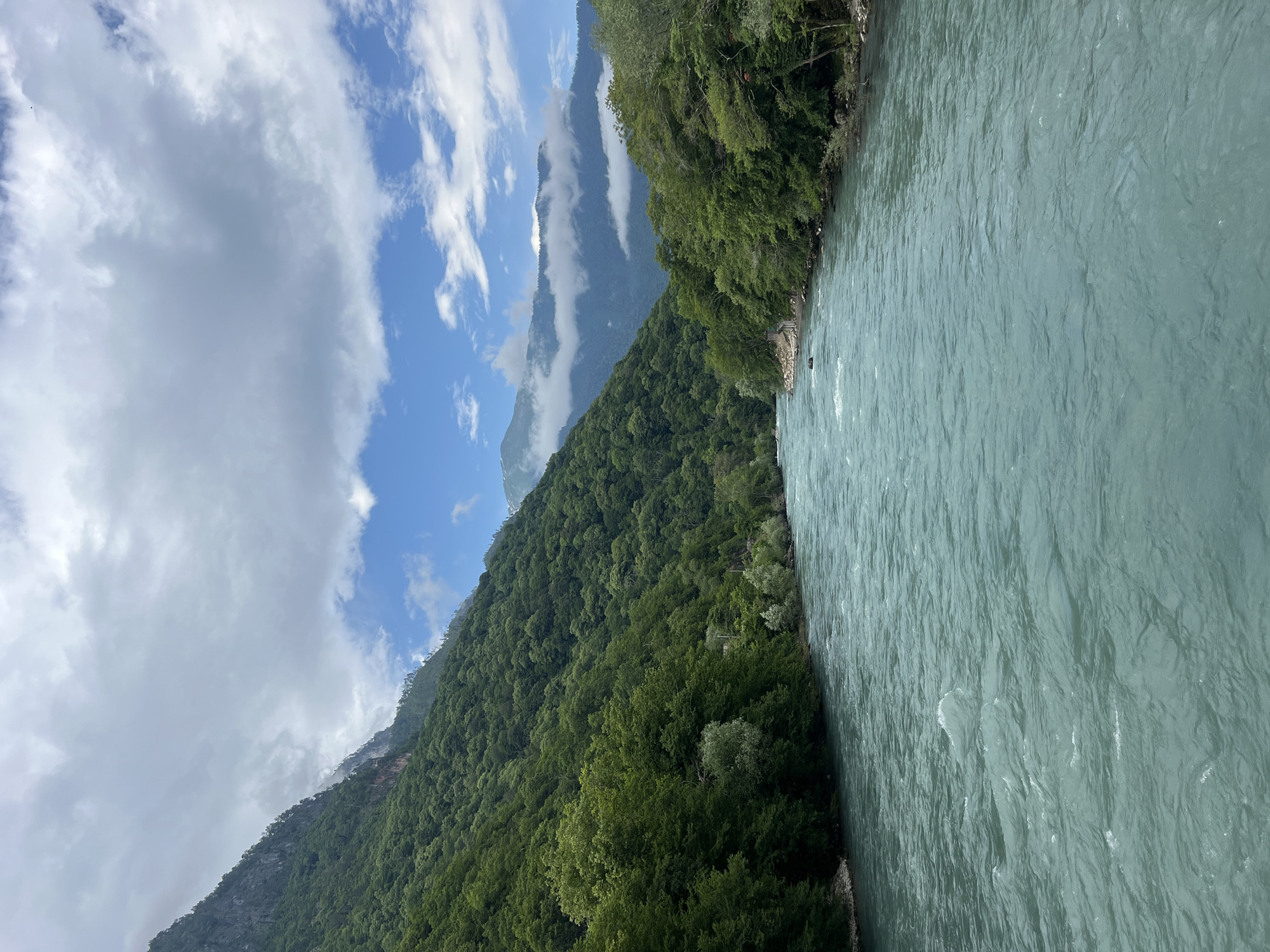 The image shows a wide, turquoise river flowing alongside a steep, heavily forested mountain. The mountain is covered in lush green trees, and its slope rises dramatically from the riverbank. In the background, taller, more rugged mountains are visible, some shrouded in wisps of white clouds. The sky is a mix of bright blue and dramatic, fast-moving clouds, suggesting dynamic weather. The river has a textured surface with ripples and small waves, indicating a current. The overall scene is one of natural beauty and rugged wilderness, likely in a mountainous region. The orientation of the image is rotated 90 degrees counter-clockwise, with the river on the right and the mountains on the left.