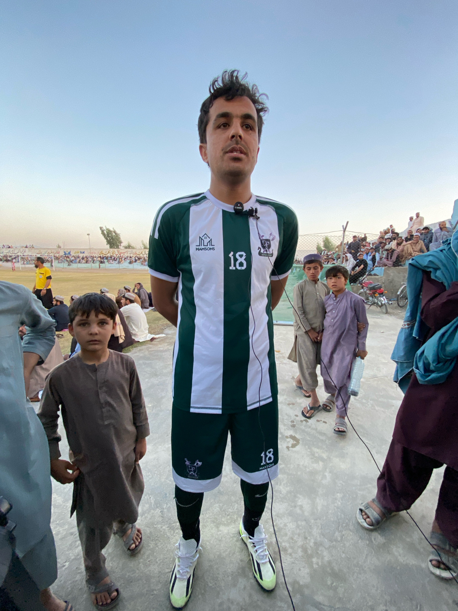 In Spin Boldak, Afghanistan, a young man wearing a green and white striped soccer jersey with the number 18 and the brand "MAMSONS" is the central focus of the image. He has a microphone clipped to his shirt and appears to be speaking. He is standing on a paved surface. In front of him, a young boy in a brown traditional outfit is looking directly at the camera. To the right of the central figure, two other boys are visible, one in a purple traditional outfit holding a water bottle, and another in a grey traditional outfit. Behind them, a crowd of people are gathered, some sitting on bleachers, suggesting a sporting event or public gathering. A motorcycle is parked in the background. The sky is clear and light blue, indicating daytime. The environment appears to be an outdoor stadium or open area.