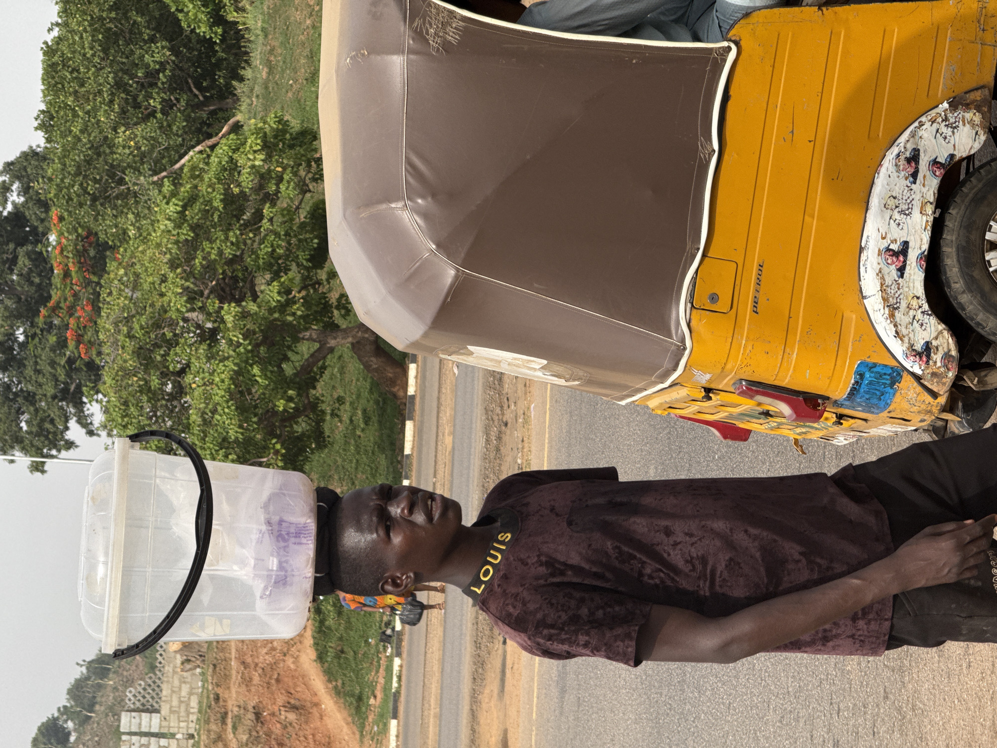 This image captures a roadside scene in Abuja, Nigeria, featuring a young man and a yellow three-wheeled vehicle.
The primary subject is a dark-skinned young man, viewed from the chest up, standing by the side of a paved road. He is carrying a large, clear plastic container with a black handle balanced on his head. He wears a dark maroon/brown short-sleeved shirt with a velvety texture and dark trousers or shorts. The black collar of his shirt prominently displays the text "LOUIS" in yellow lettering. His facial expression is neutral, and he appears to be looking slightly upwards or ahead.
To his right, partially visible, is a yellow three-wheeled vehicle, commonly known as a Keke NAPEP or auto-rickshaw in Nigeria, with a brown canvas canopy. The side of the vehicle clearly shows the word "PETROL" printed on it. The rear fender of the vehicle is decorated with white and patterned designs, including multiple small images of faces or figures. Another person's legs are visible inside the vehicle, suggesting it is occupied.
The setting appears to be outdoors during daytime, indicated by the bright natural lighting. Lush green trees and vegetation dominate the background, with some trees showing clusters of red flowers. The road is asphalt, and to the left of the man, some grey concrete blocks or construction materials are visible. The overall impression is that of a bustling or semi-urban roadside environment in Abuja.