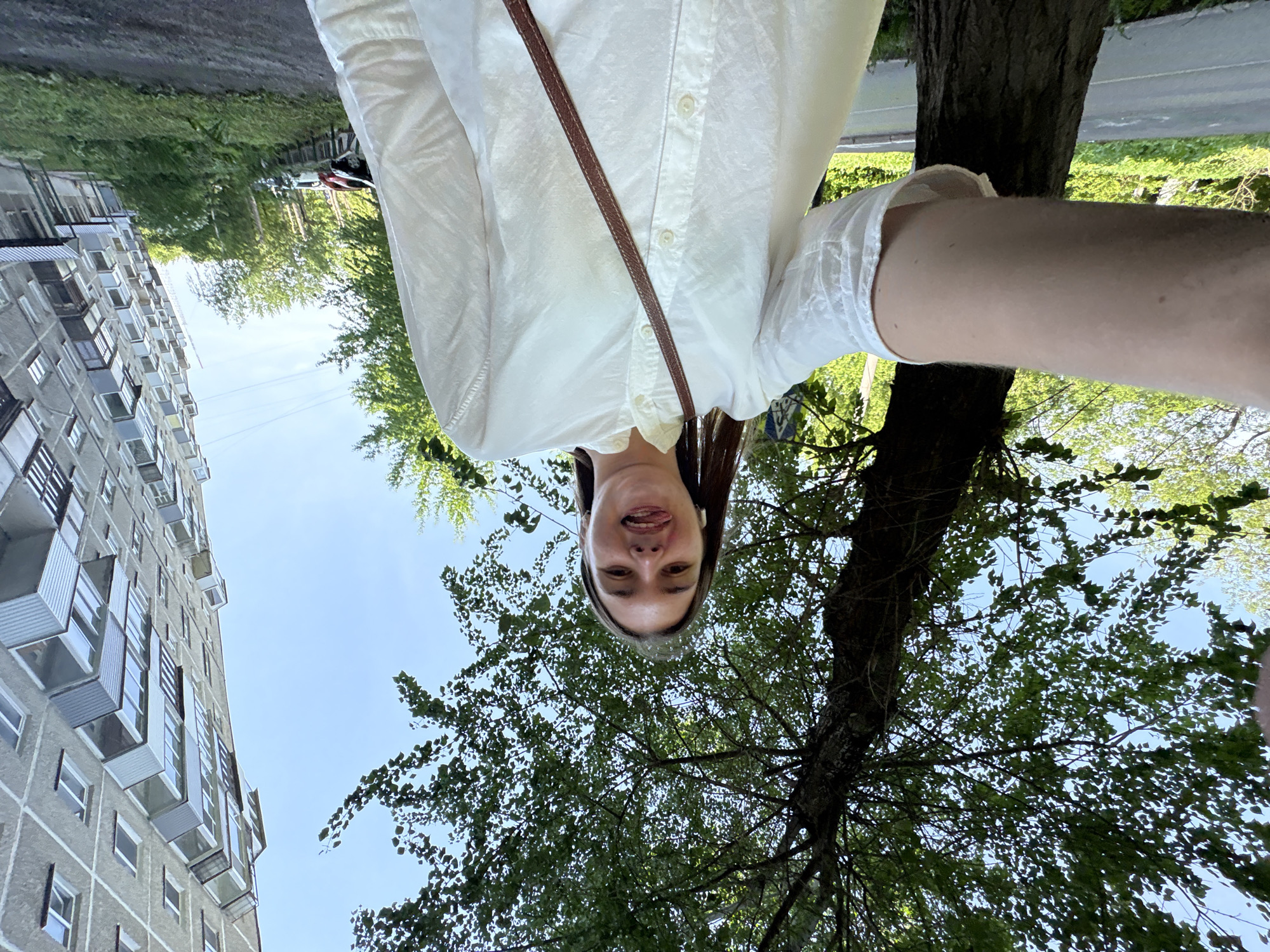 A selfie depicts a woman in Perm, Russia, standing outdoors. She is wearing a white button-down shirt with a brown crossbody strap. The building has grey exteriors with windows and protruding balconies. A tree with green leaves is on the right side of the image. The sky is blue.