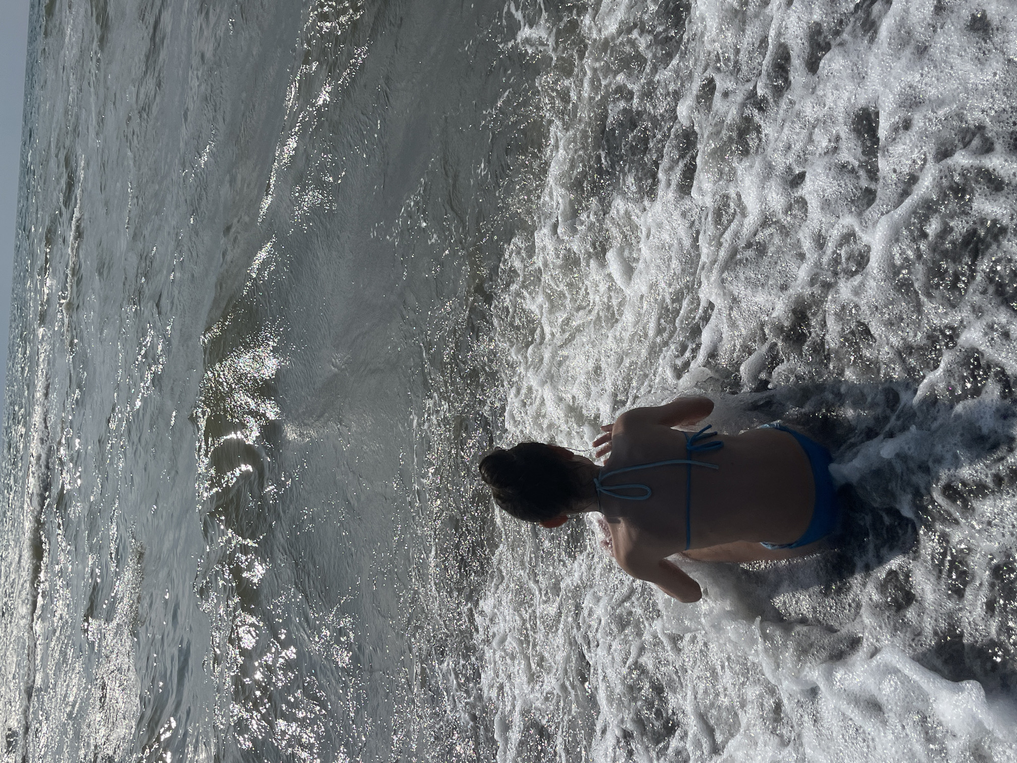 A person, viewed from behind, sits in the shallow water of the sea at Ureki, Georgia. The individual is wearing a blue bikini and has their hair pulled up in a bun. Waves are breaking around them, creating white foam. The water is choppy and reflects the sunlight, creating a shimmering effect. The horizon is visible in the distance, with a hazy sky.