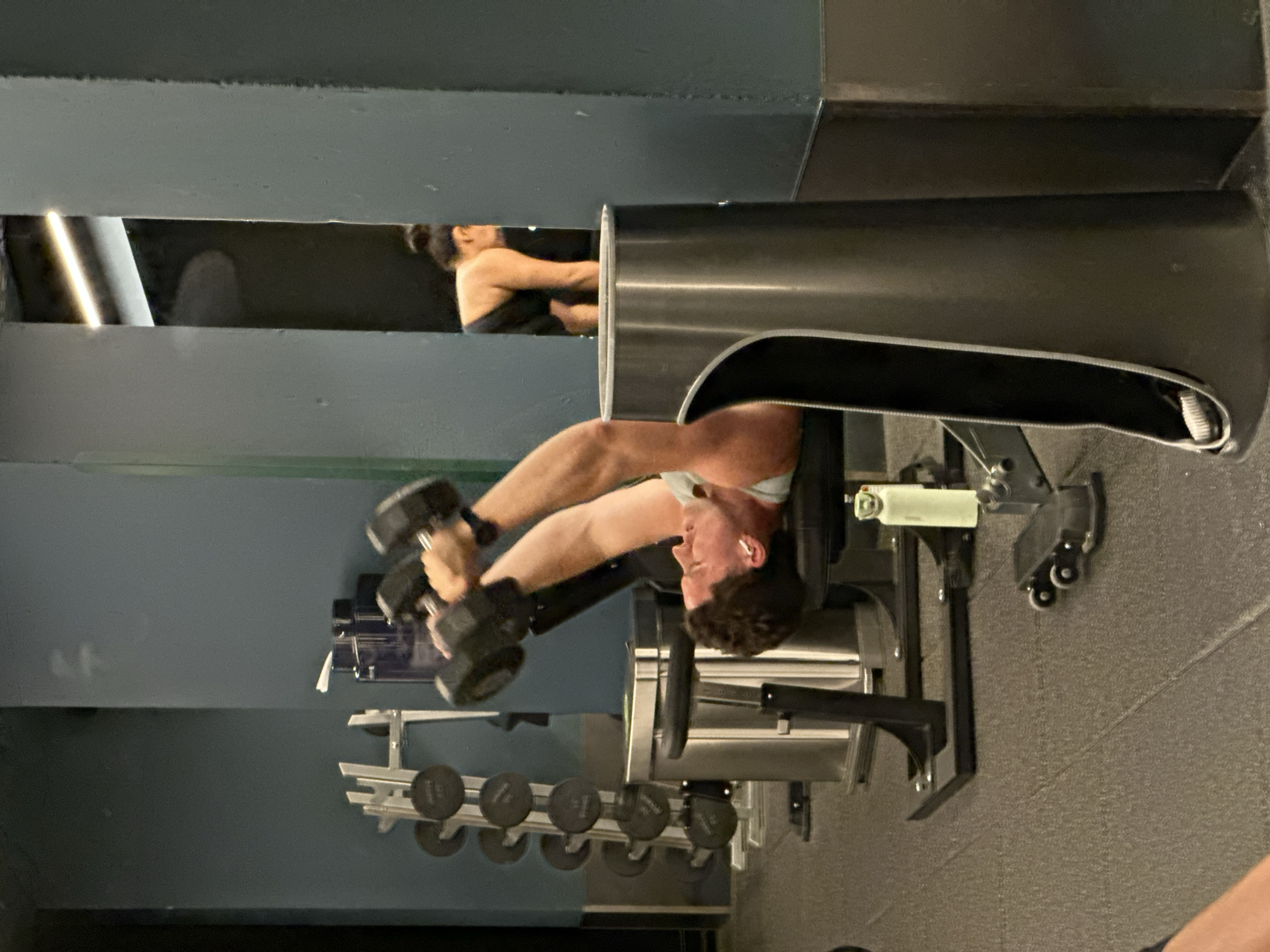 The image depicts an interior scene within a gym or fitness center, characterized by dark walls and artificial lighting. The primary subject is a man, positioned upside down on what appears to be an inversion table or a hyperextension bench, engaging in an exercise. He holds a black hexagonal dumbbell in each hand, extending his arms. He has short, curly dark hair, wears a light-colored top (possibly light blue or white) and light shorts, and has white wireless earbuds in his ears, suggesting he is listening to music or audio. His expression appears focused on his activity.
In the upper left quadrant of the image, seen through a rectangular opening in a dark wall, a second individual, a woman, is partially visible. She is seen from the back or side, wearing a dark athletic top, and appears to be in the process of exercising or stretching, though her specific action is unclear.
The setting includes various gym equipment. Below the man, a rack of black hexagonal dumbbells of different weights is visible, neatly arranged. The floor is a textured grey, likely a rubberized gym surface. The equipment the man is using is robust, featuring metallic and dark components, with small wheels visible on its base. There are no explicit visual cues within the image itself to confirm the London, United Kingdom location, but this context is provided. No visible text can be clearly discerned on any objects or signage. The time of day is not indicated due to the indoor setting and artificial light.