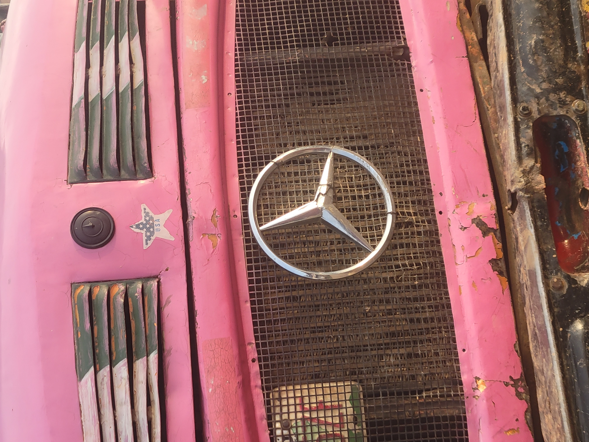 The image shows a close-up of the front grille of a vehicle, painted pink. The paint is peeling and cracked in several places, revealing underlying layers. A silver Mercedes-Benz emblem is prominently displayed in the center of the grille. To the left of the grille are vertical slats, painted with alternating green and white stripes. Above these slats is a circular black object and a sticker with a star and "USA" printed on it. To the right of the grille, there is a dark, rusted metal component with visible screws. The background is out of focus, but appears to be a gritty, urban environment. The location is Unknown, Unknown.