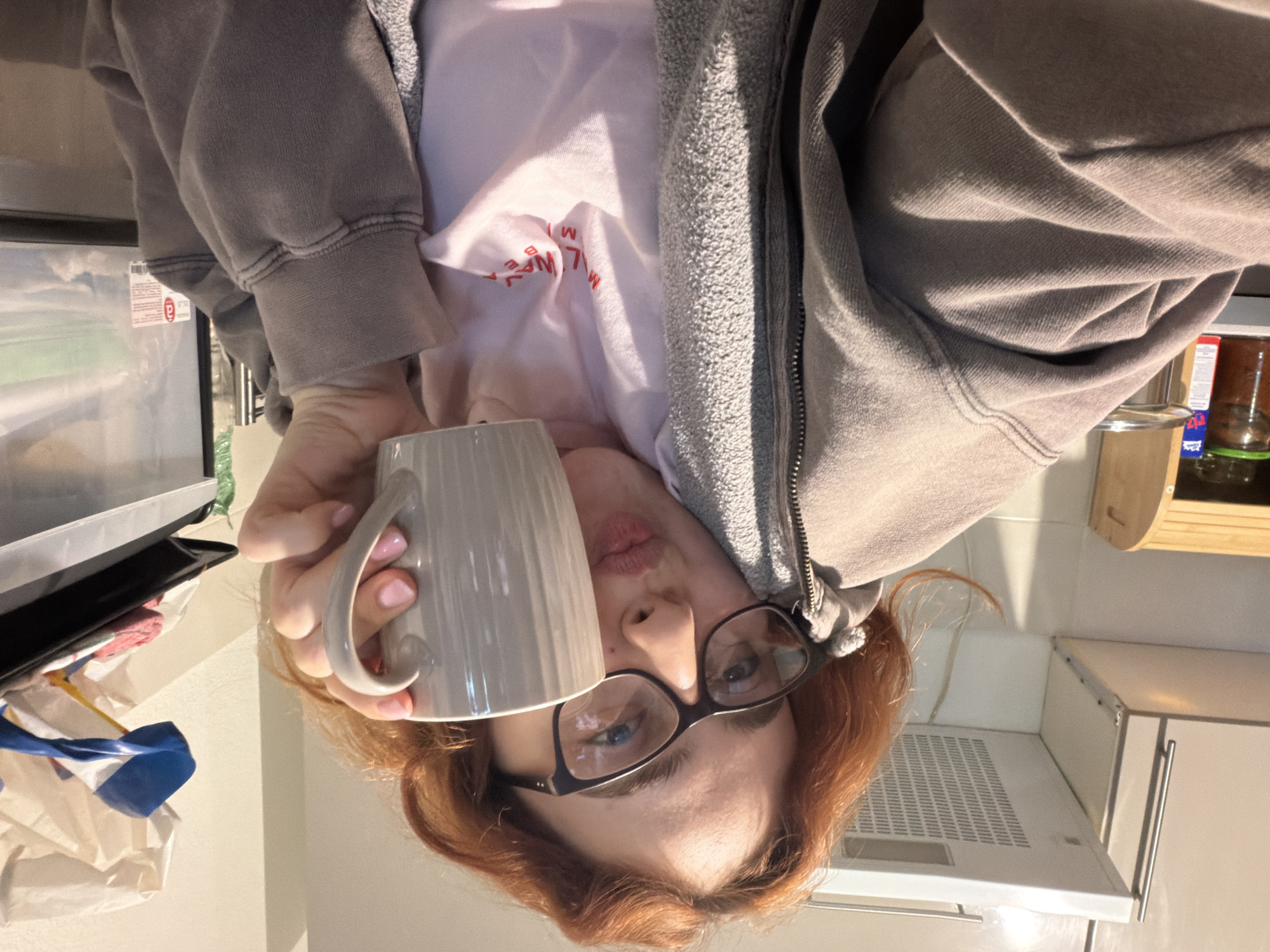 A person with reddish-brown hair and glasses is holding a light gray mug in their right hand, with their lips pursed in a kiss. They are wearing a light pink t-shirt and a gray hooded sweatshirt. The background shows a kitchen area with cabinets, a range hood, a wooden bread box containing jars, and a plastic drawer unit filled with various items. The scene is indoors and appears to be a residential kitchen. The location of Strasbourg, France is not evident from the image.