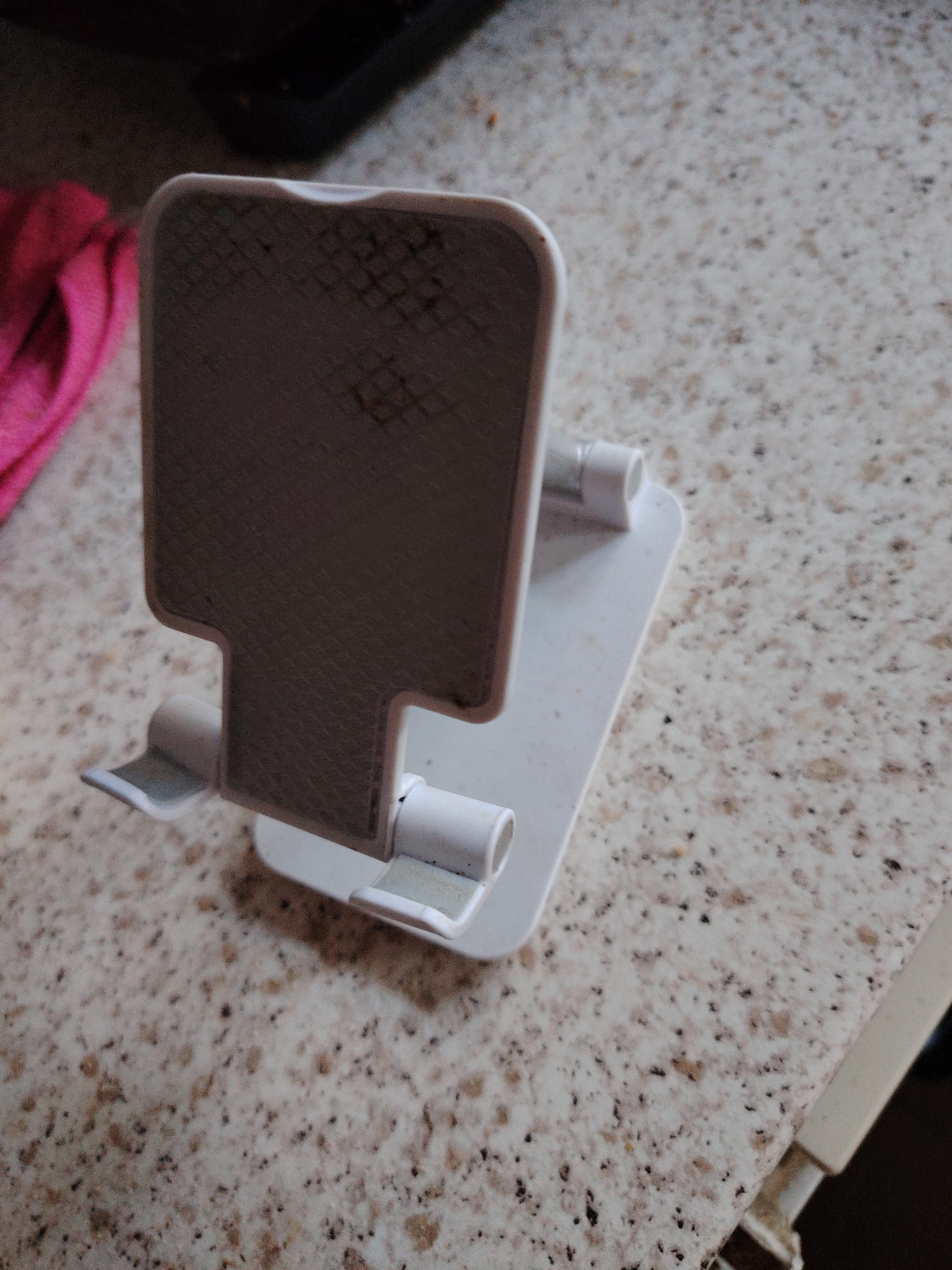 A white plastic mobile phone stand is positioned on a speckled countertop. The stand has a textured backplate and two small prongs at the bottom to hold a phone. A pink cloth is visible in the background to the left of the stand. The countertop is light-colored with dark speckles. The scene is indoors, likely in a kitchen or similar domestic environment. The specific location in Huntingdonshire, United Kingdom is not identifiable from the image.