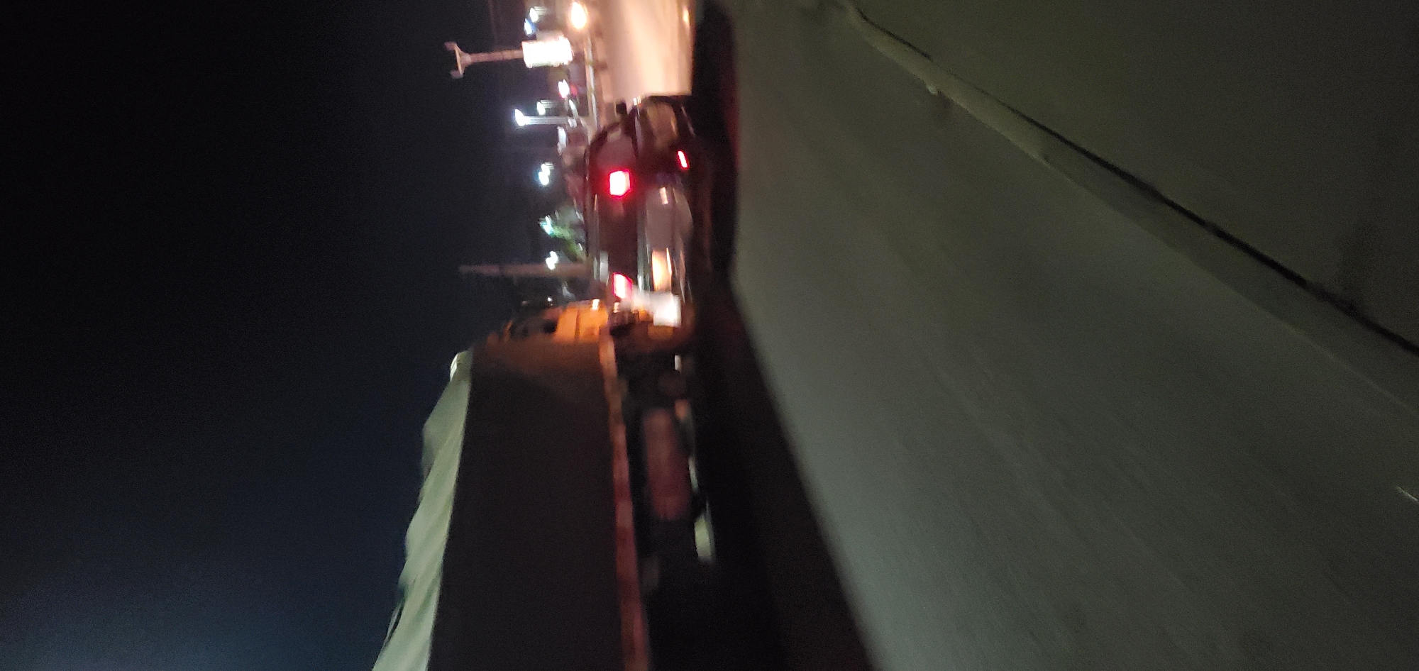 The image presents a nighttime street scene in Gura Top, Nigeria. A large, dark-colored cargo truck with a light-colored, possibly canvas, cover on its bed is visible on the left side, appearing to move from left to right. Its cabin shows warm yellow illumination. To the right of the truck, a passenger vehicle, likely a car or SUV, is also in motion, displaying illuminated red taillights. Both vehicles and the immediate foreground exhibit motion blur. The road surface is dark and appears to be asphalt or concrete, with a distinct curb line present along the lower right edge. The background consists of a dark night sky with various distant, blurred artificial light sources, suggesting streetlights or other urban illumination. No individuals are distinctly visible.